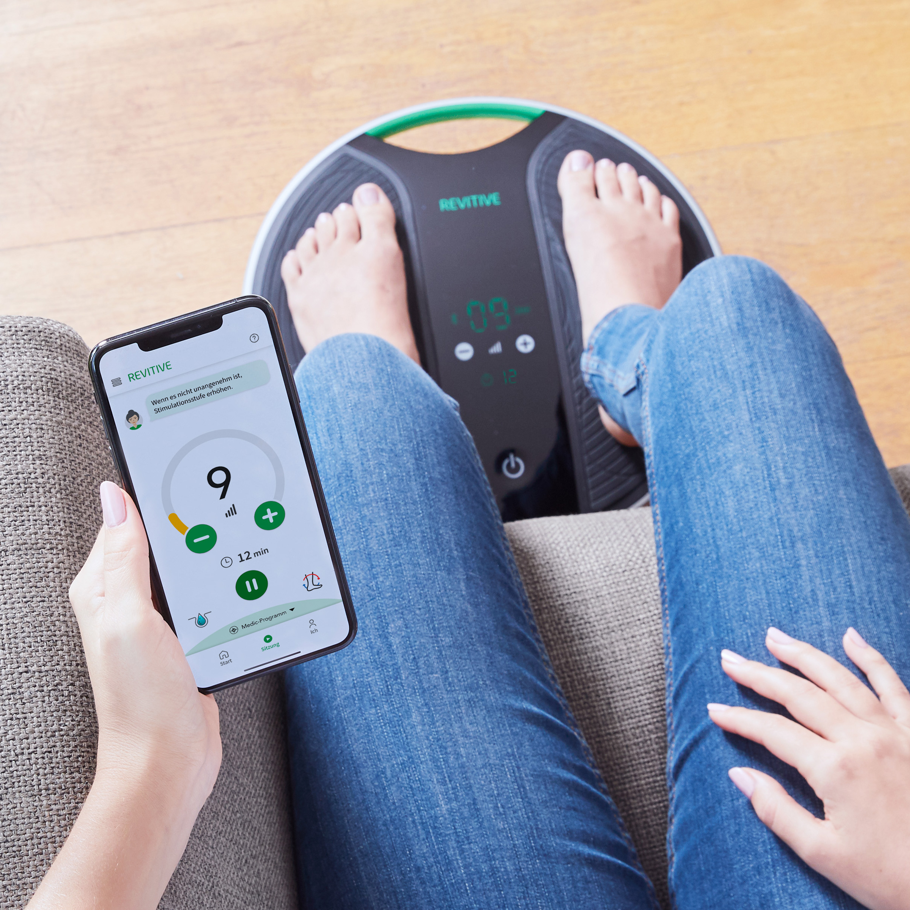 Person nutzt Smartphone und steht auf Revitive Fußmassagegerät. Jeans und graues Sofa.