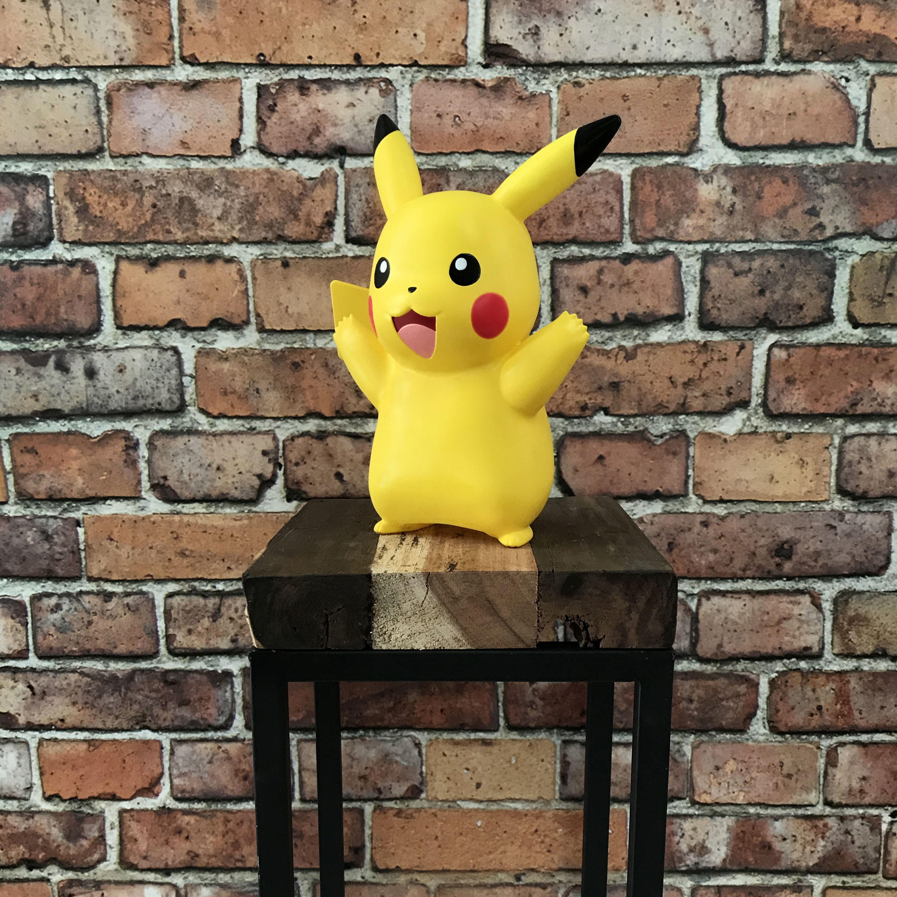 Statua di Pikachu giallo su un supporto nero. Sfondo di muro di mattoni rossi. Braccia alzate.