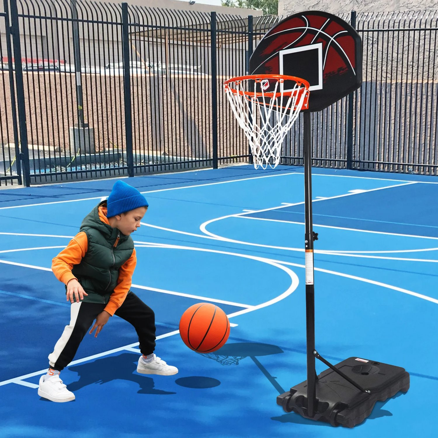 Ein Junge dribbelt einen Basketball in Richtung eines Korbs auf einem blauen Platz. Schwarzer Zaun im Hintergrund.
