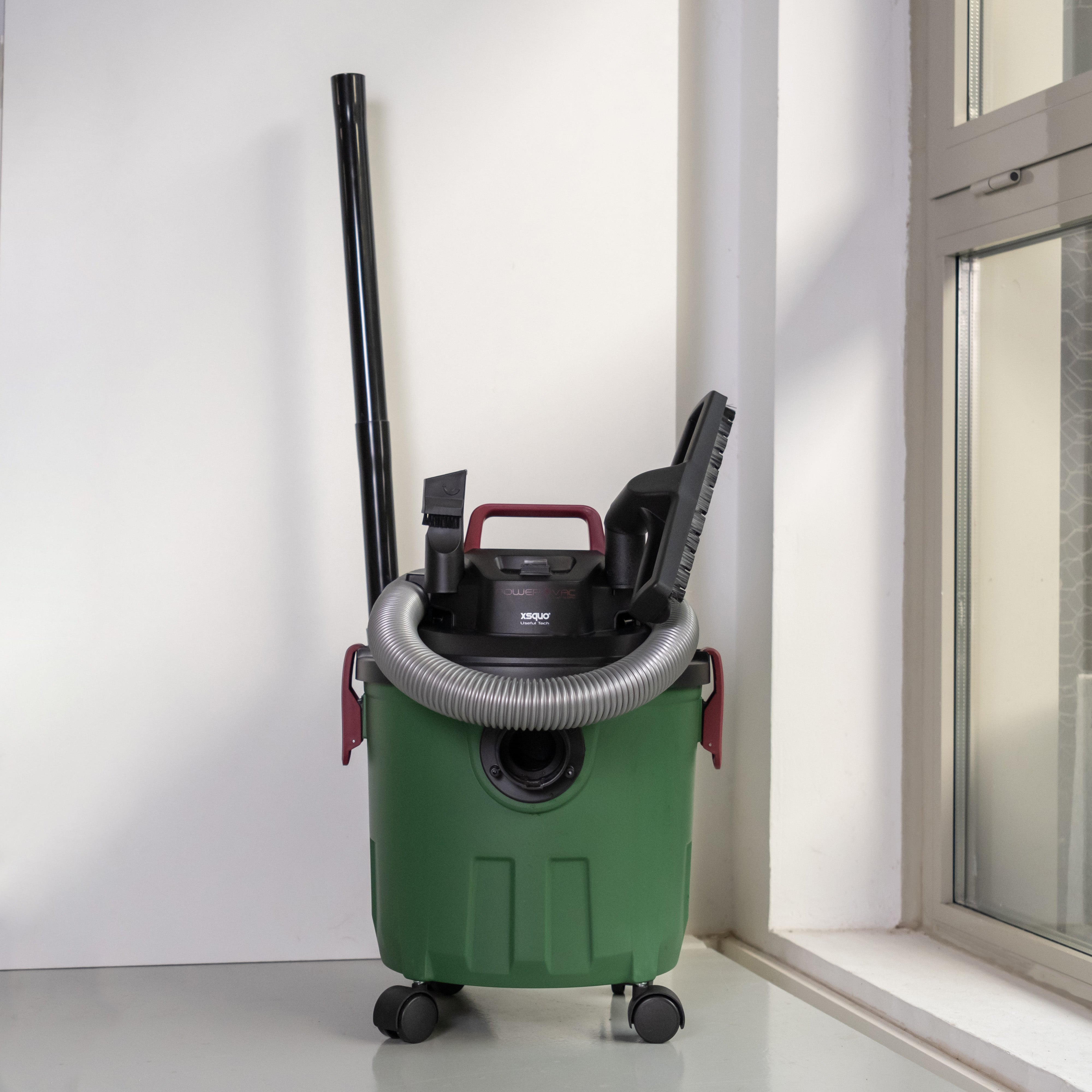 Aspiradora para líquidos y sólidos verde y negra con accesorios, cerca de una pared blanca y una ventana.