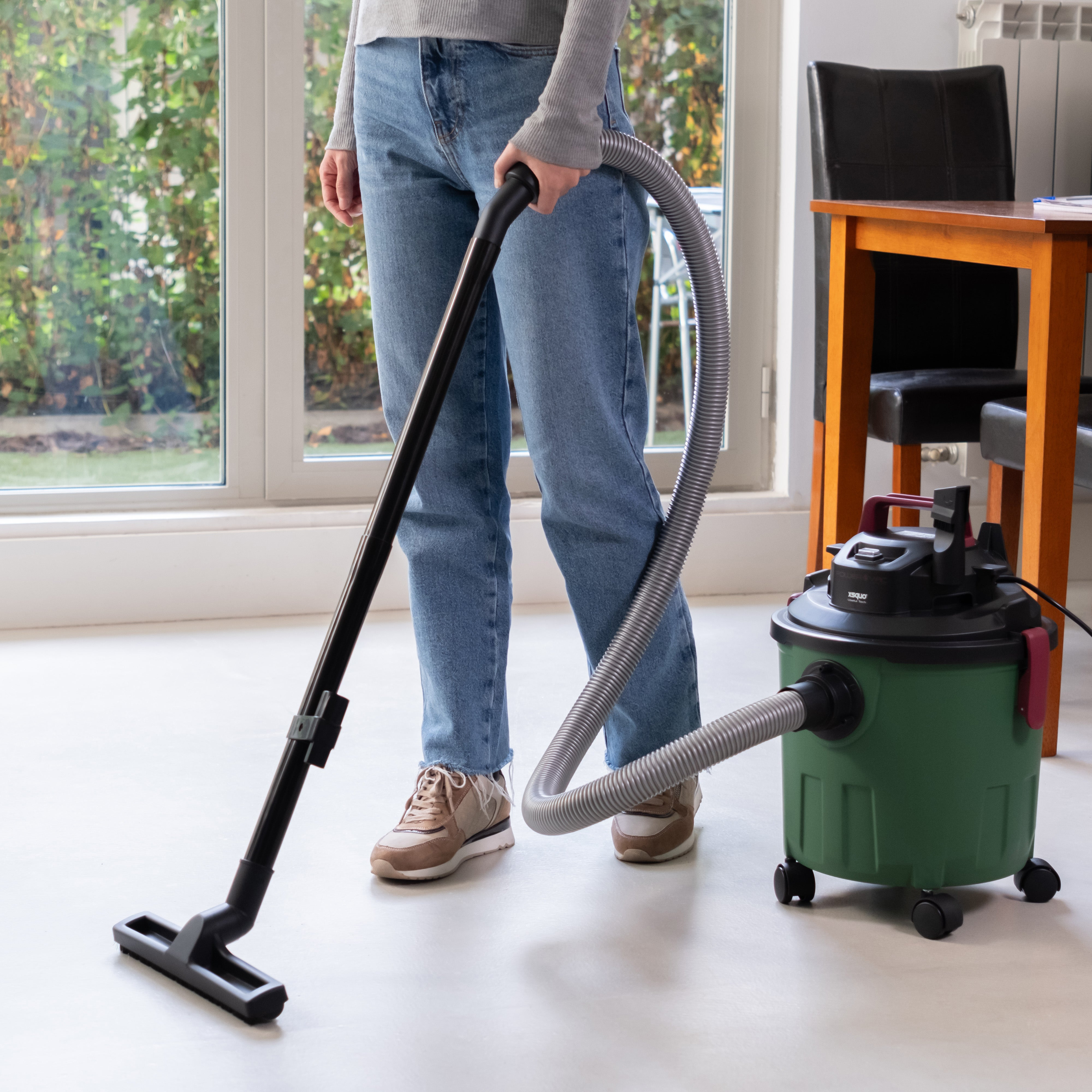 Persona aspirando un suelo blanco en una habitación con una aspiradora verde y grandes ventanas.