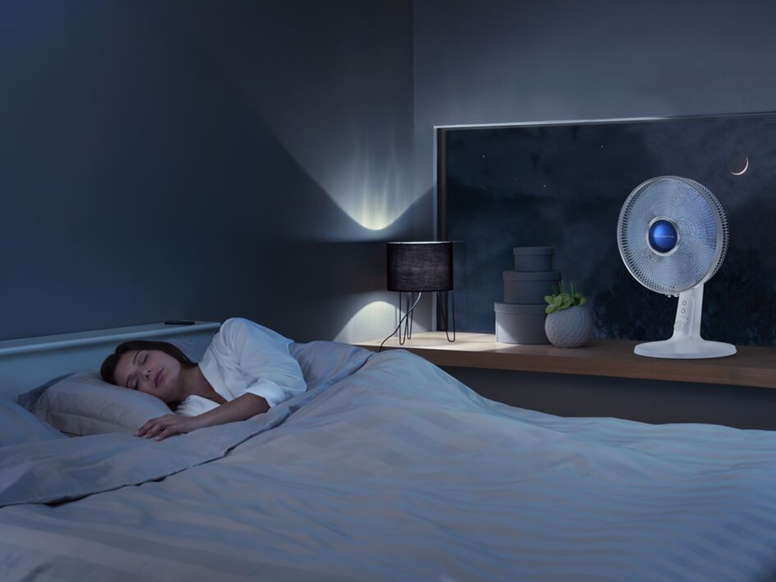 Kobieta śpiąca w ciemnej sypialni. Wentylator stoi na półce blisko okna.