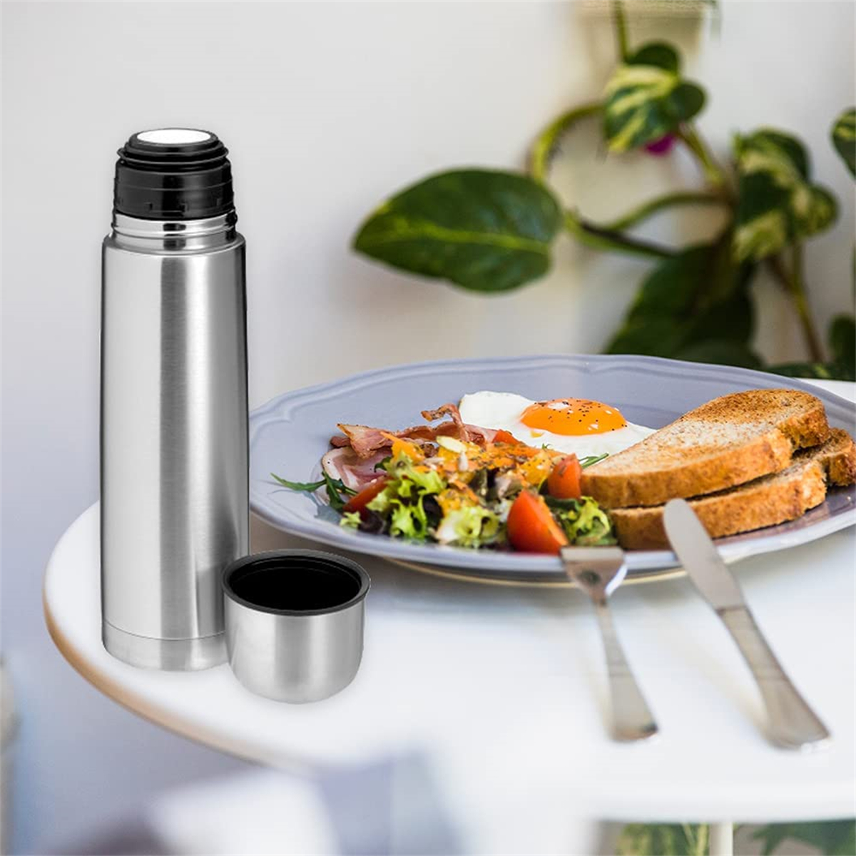 Eine Edelstahl-Thermoskanne mit schwarzem Deckel und einer Tasse daneben, neben einem Teller mit Essen.