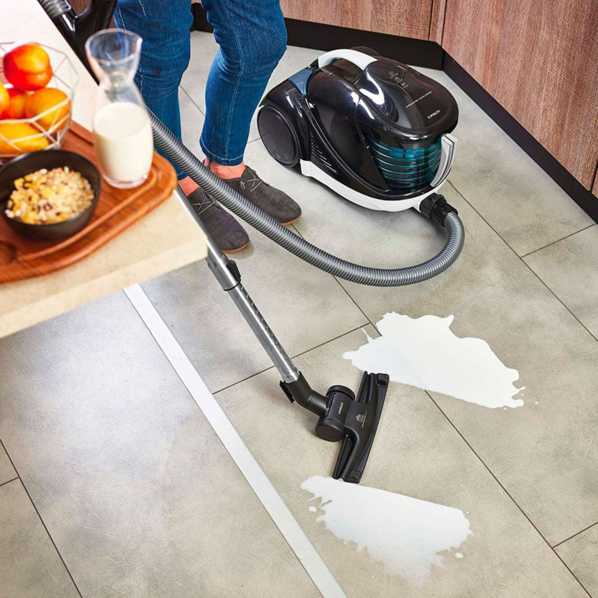 Kobieta odkurza rozlane mleko na szarej podłodze z płytek, miska z jedzeniem i dzbanek na blacie.