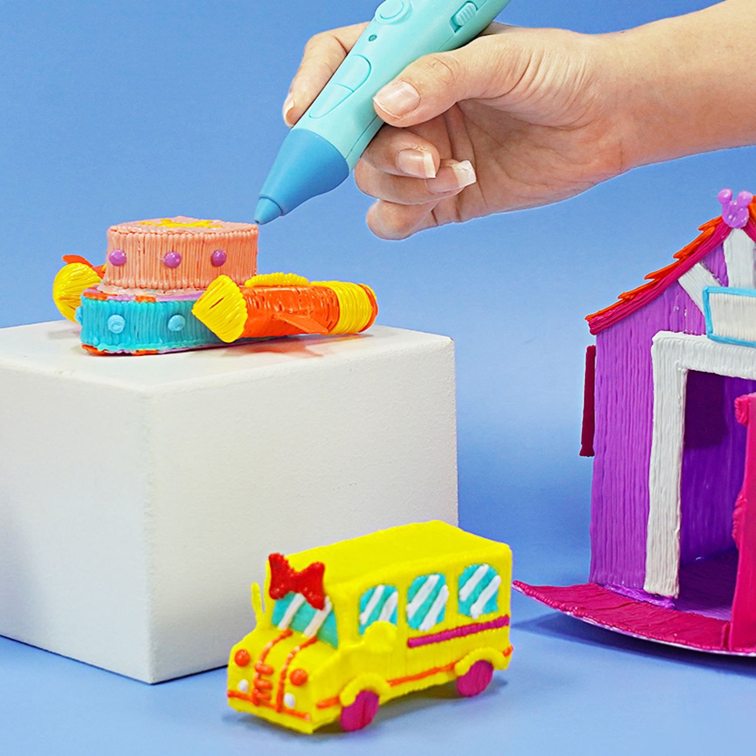 Eine Hand benutzt einen blauen 3D-Stift, um bunte Modelle auf einem weißen Block zu erstellen. Enthält einen Schulbus und Kuchen.