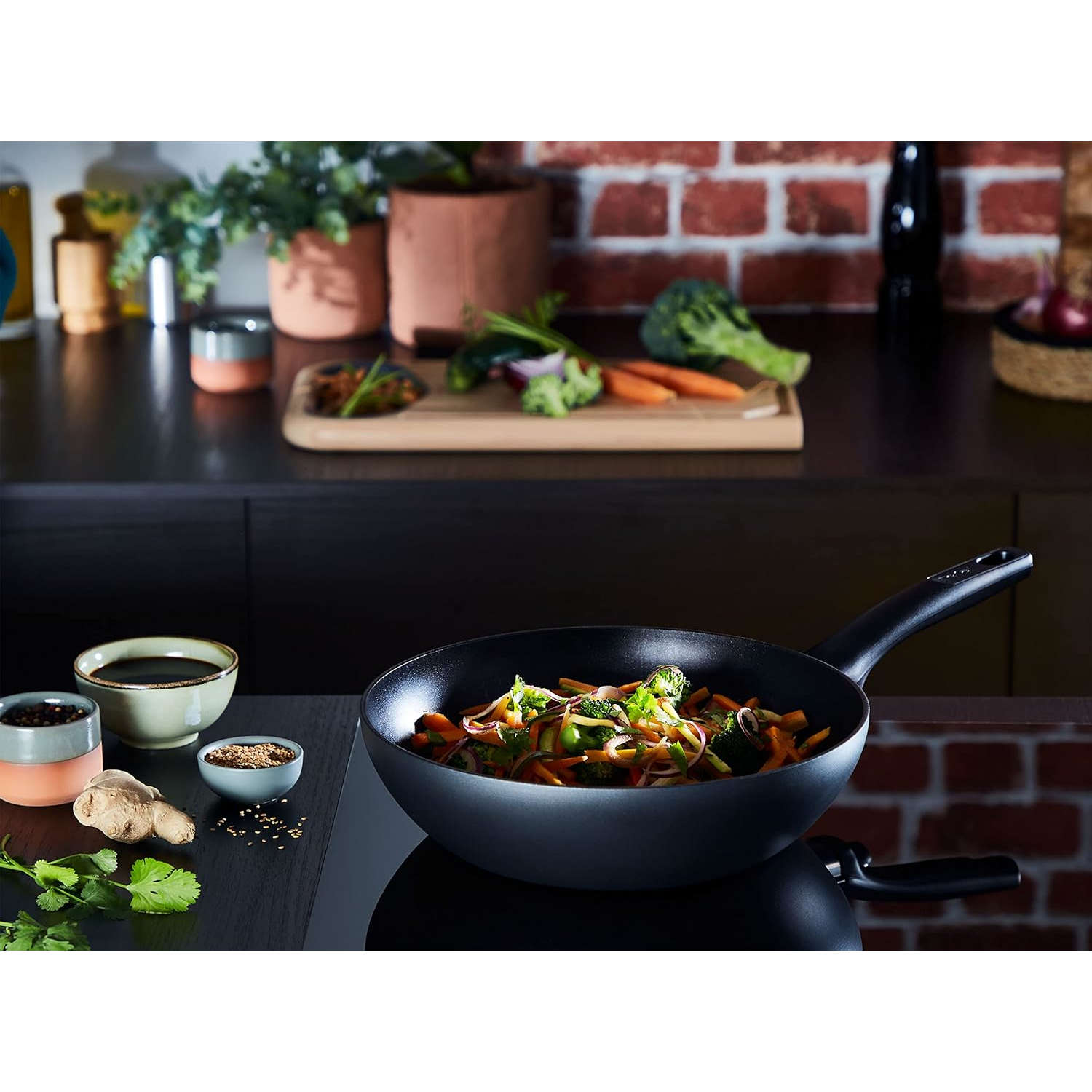 Ein Wok mit gebratenem Gemüse auf einem schwarzen Herd, mit Zutaten und Pflanzen im Hintergrund.
