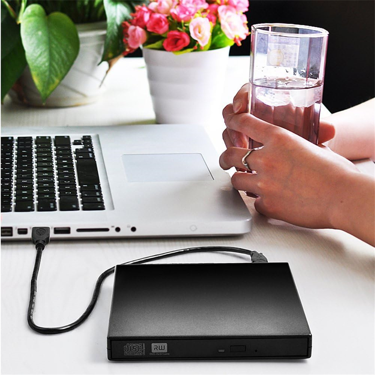Ein Laptop steht neben einem schwarzen DVD-Laufwerk. Eine Person hält ein Glas Wasser in der Nähe einer Blumenvase.