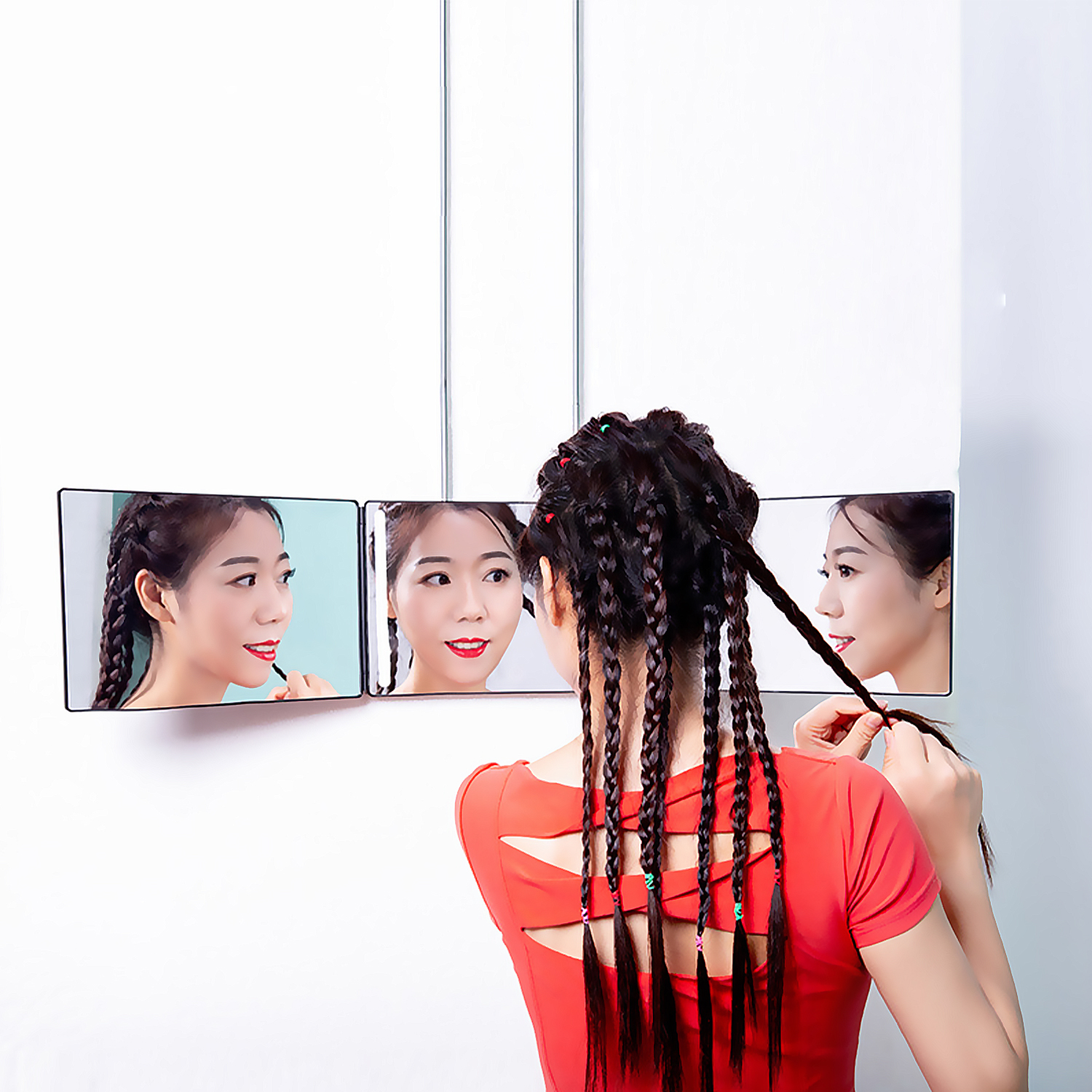 Mujer trenzando el cabello frente a tres espejos, con top rojo. Fondo de pared blanca.