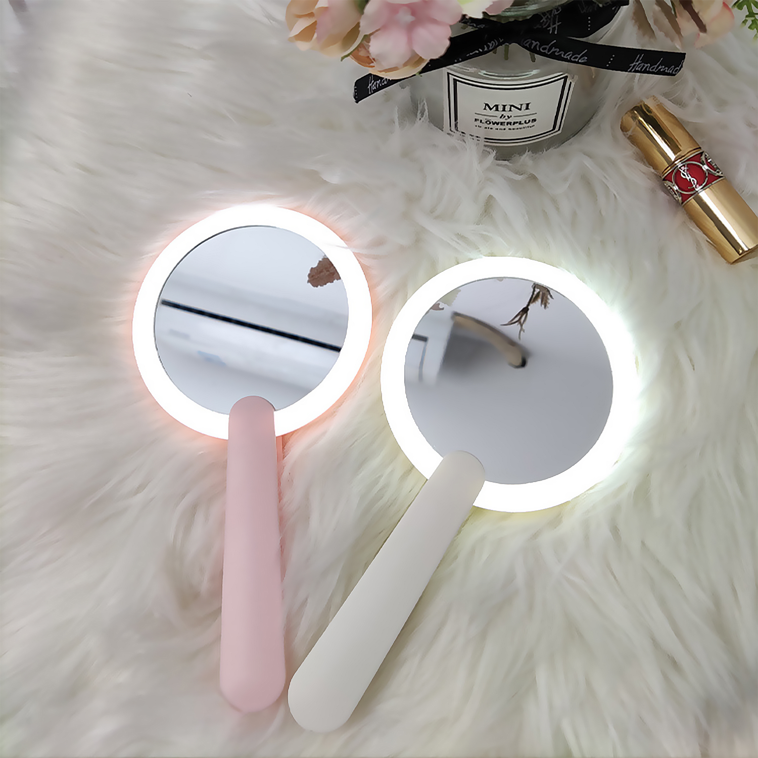 Dos espejos iluminados con asas sobre una superficie blanca y esponjosa. Lápiz labial rojo y frasco de perfume cerca.