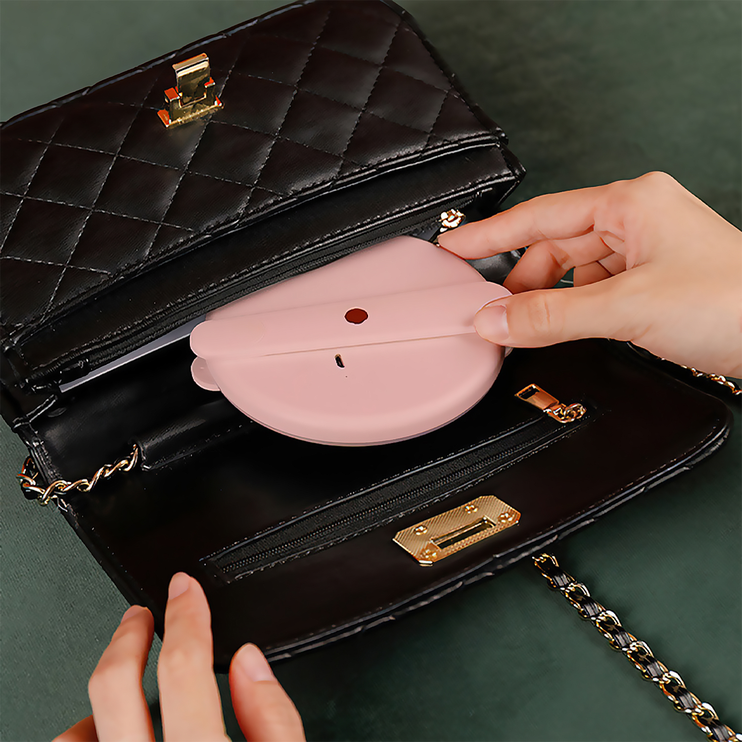 Un espejo compacto rosa, plegado, se coloca en un bolso negro con una correa de cadena dorada.