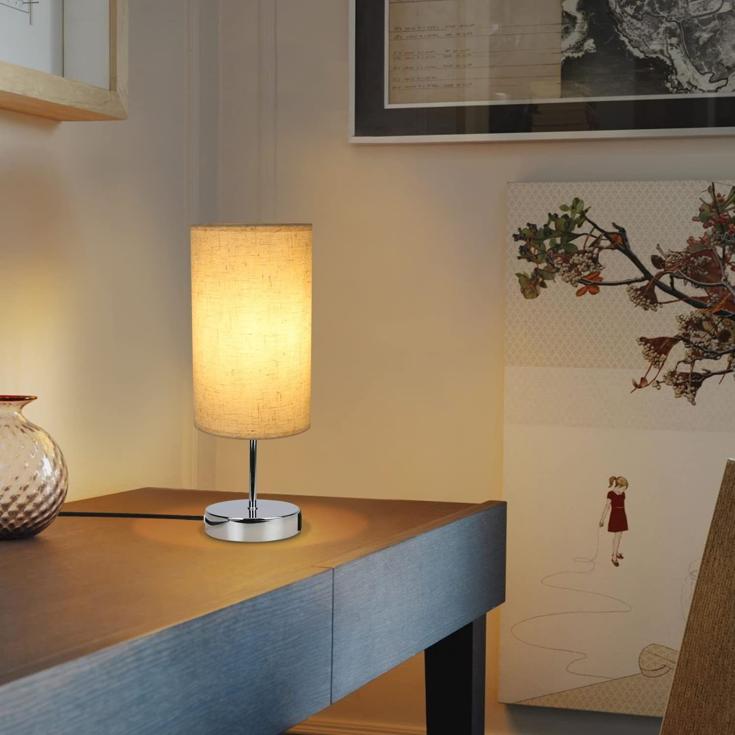 Lampa z beżowym abażurem na ciemnym drewnianym biurku, z obrazem i wazonem w tle.