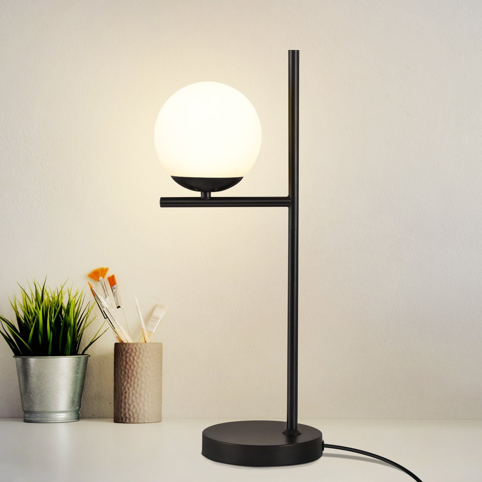 Czarna metalowa lampa biurkowa z okrągłym białym światłem i zieloną rośliną w srebrnej doniczce.