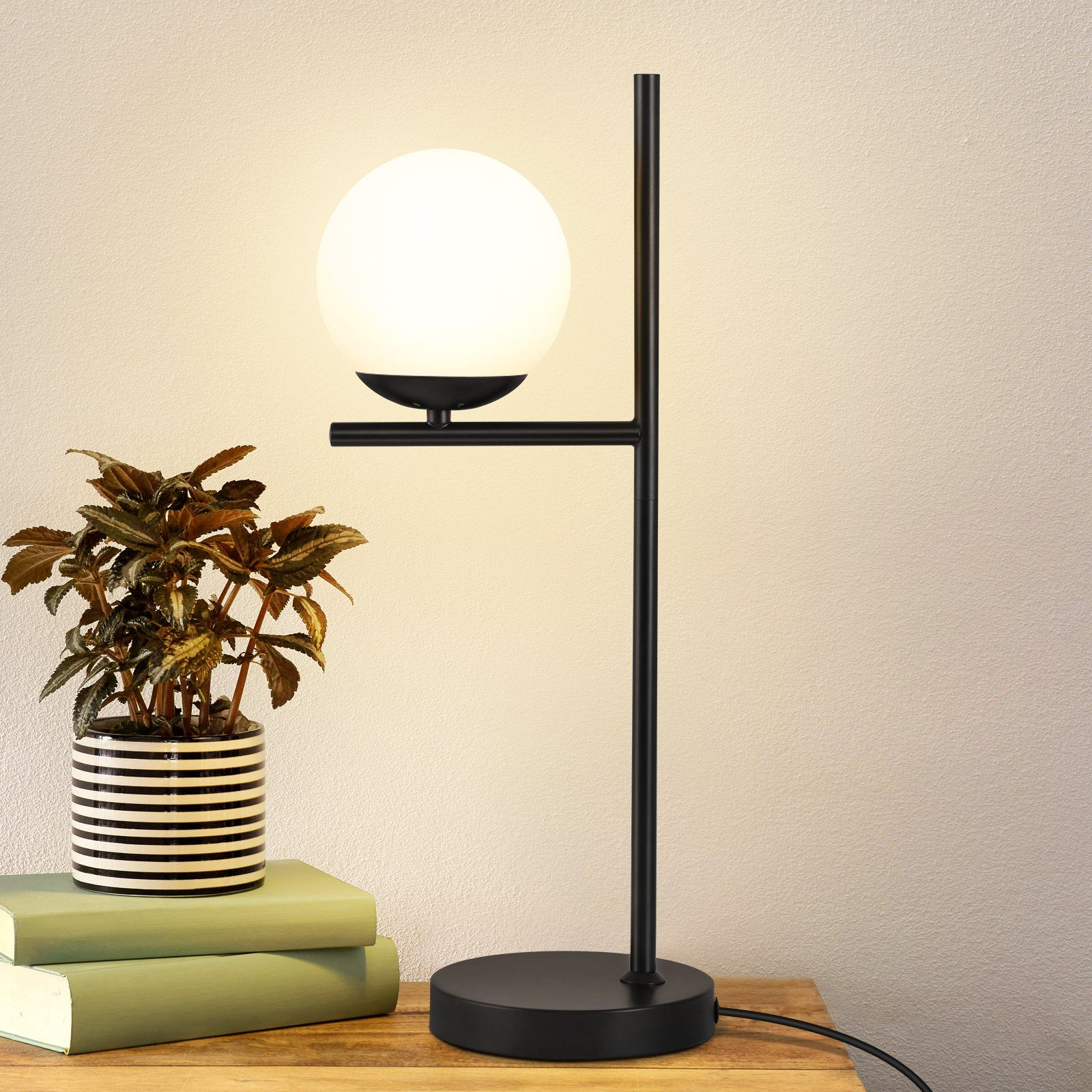 Czarna lampa z białą kulą i rośliną na drewnianym stole. Tło to ściana.