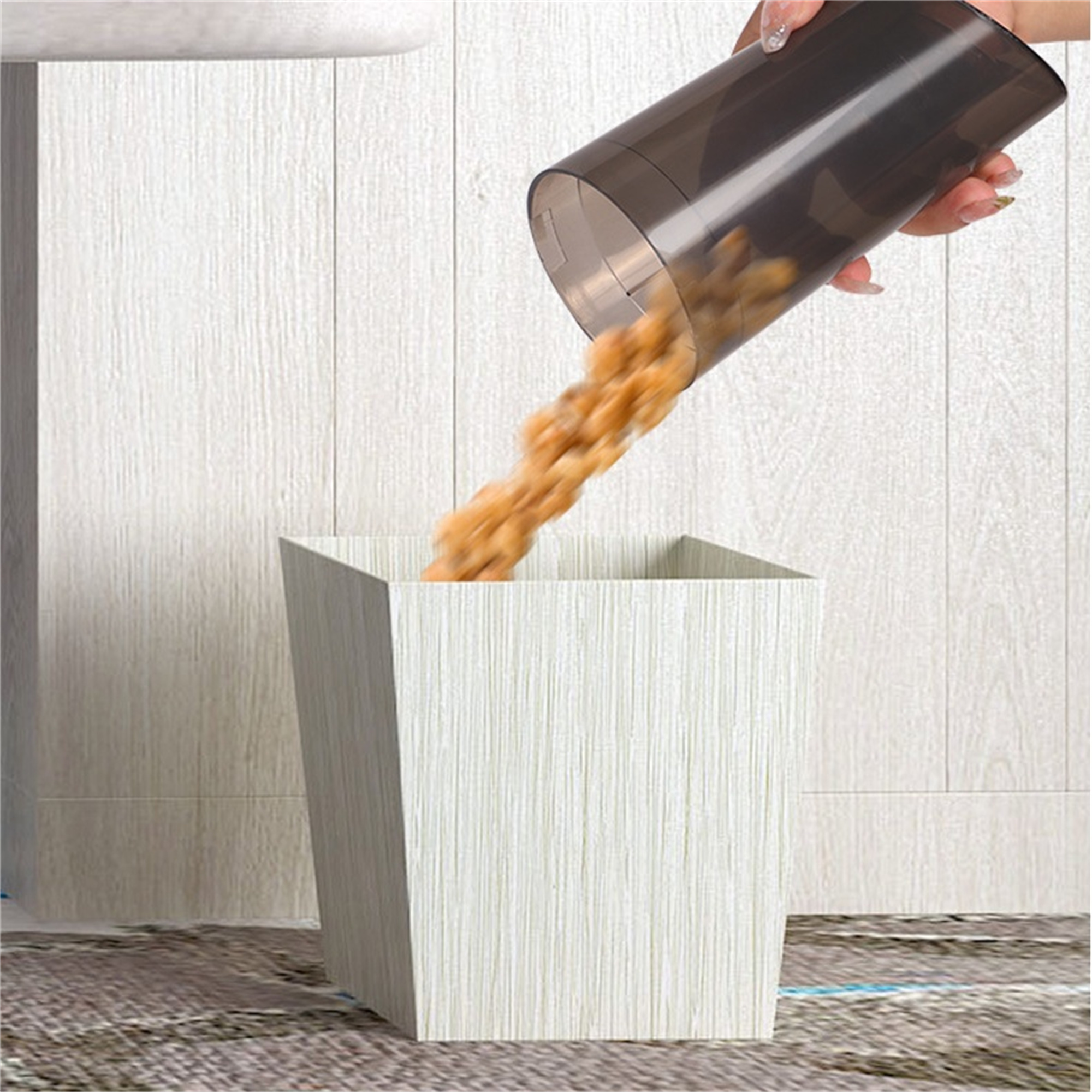 Eine Hand gießt Müsli in einen weißen, holzstrukturierten Mülleimer. Der Hintergrund ist eine helle Wand.