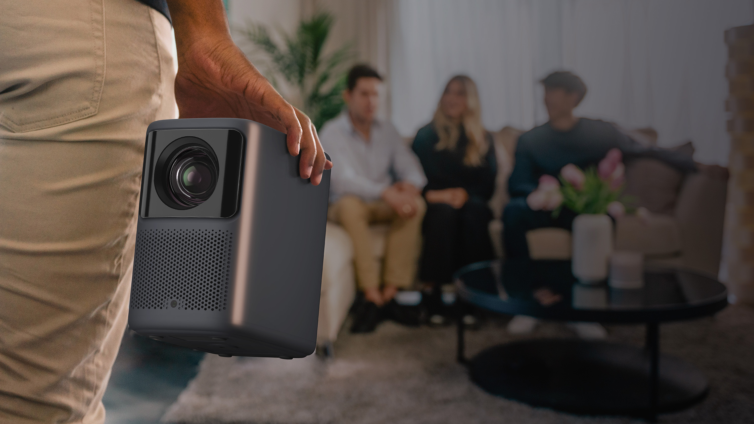 Dłoń trzyma czarny projektor. Ludzie siedzą na kanapie w rozmytym tle.