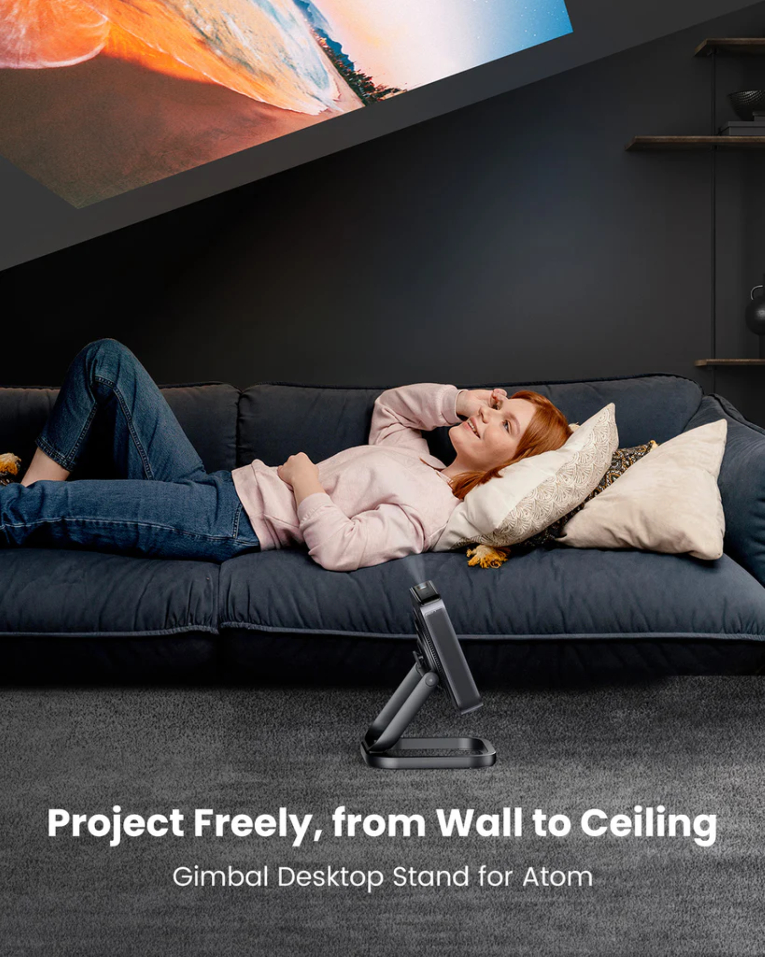 Donna sdraiata sul divano guarda una proiezione. Proiettore su un supporto. Testo: 'Project Freely, from Wall to Ceiling'.