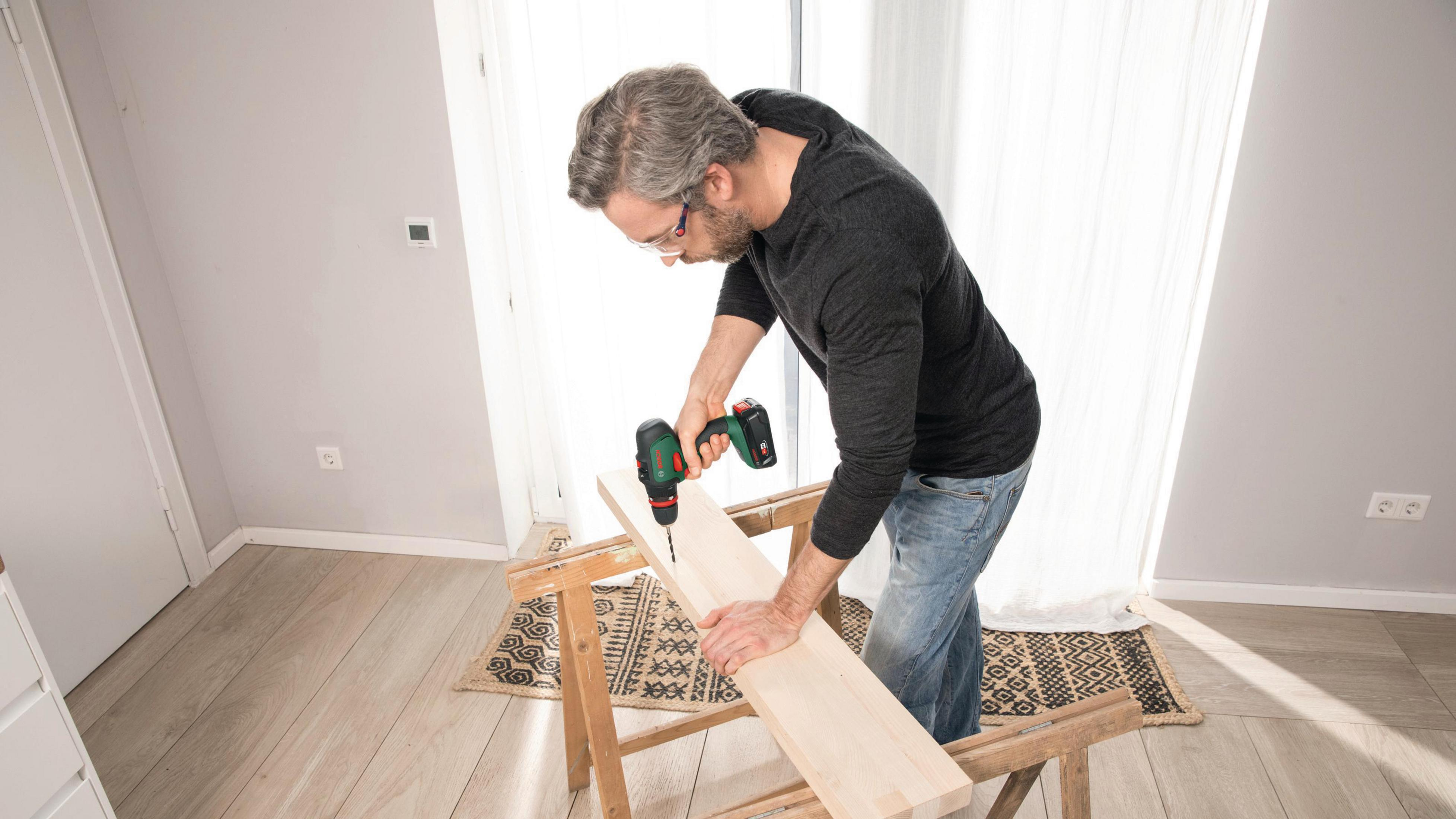 Ein Mann bohrt mit einem Akkubohrer in eine Holzplatte. Der Raum hat hellfarbige Wände.