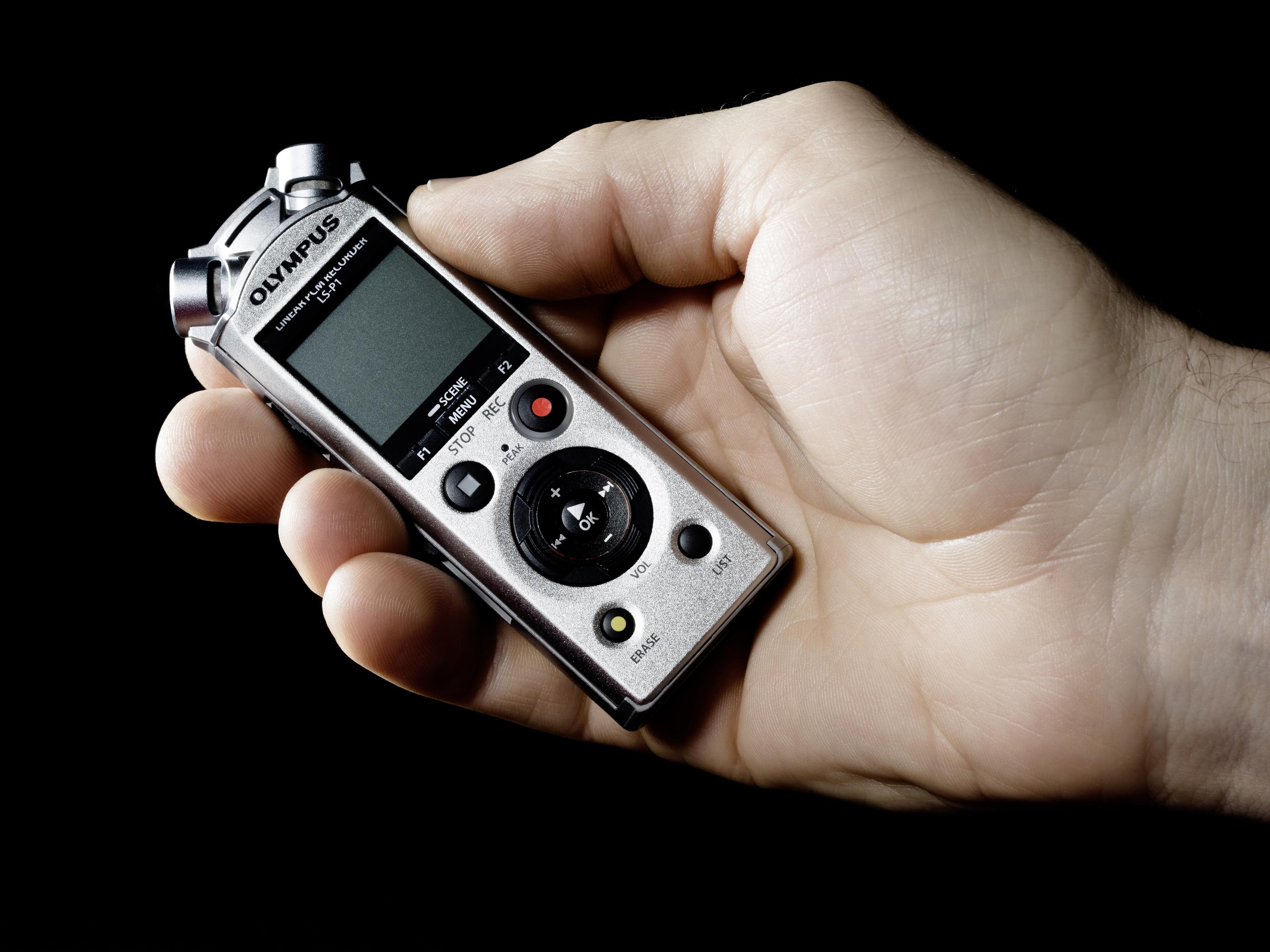 Une main tenant un enregistreur vocal numérique argenté Olympus LS-P1, sur fond noir.