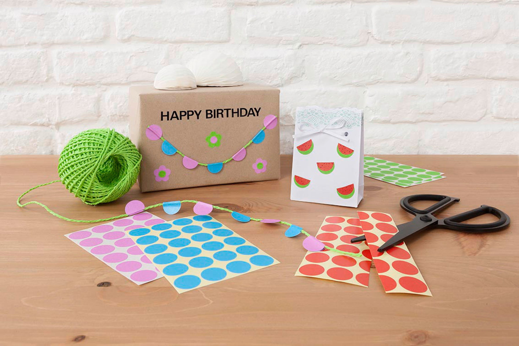 Bastelmaterialien: eine Geschenkbox, eine weiße Tüte, Schere, Aufkleber und grüne Schnur auf einem Holztisch.
