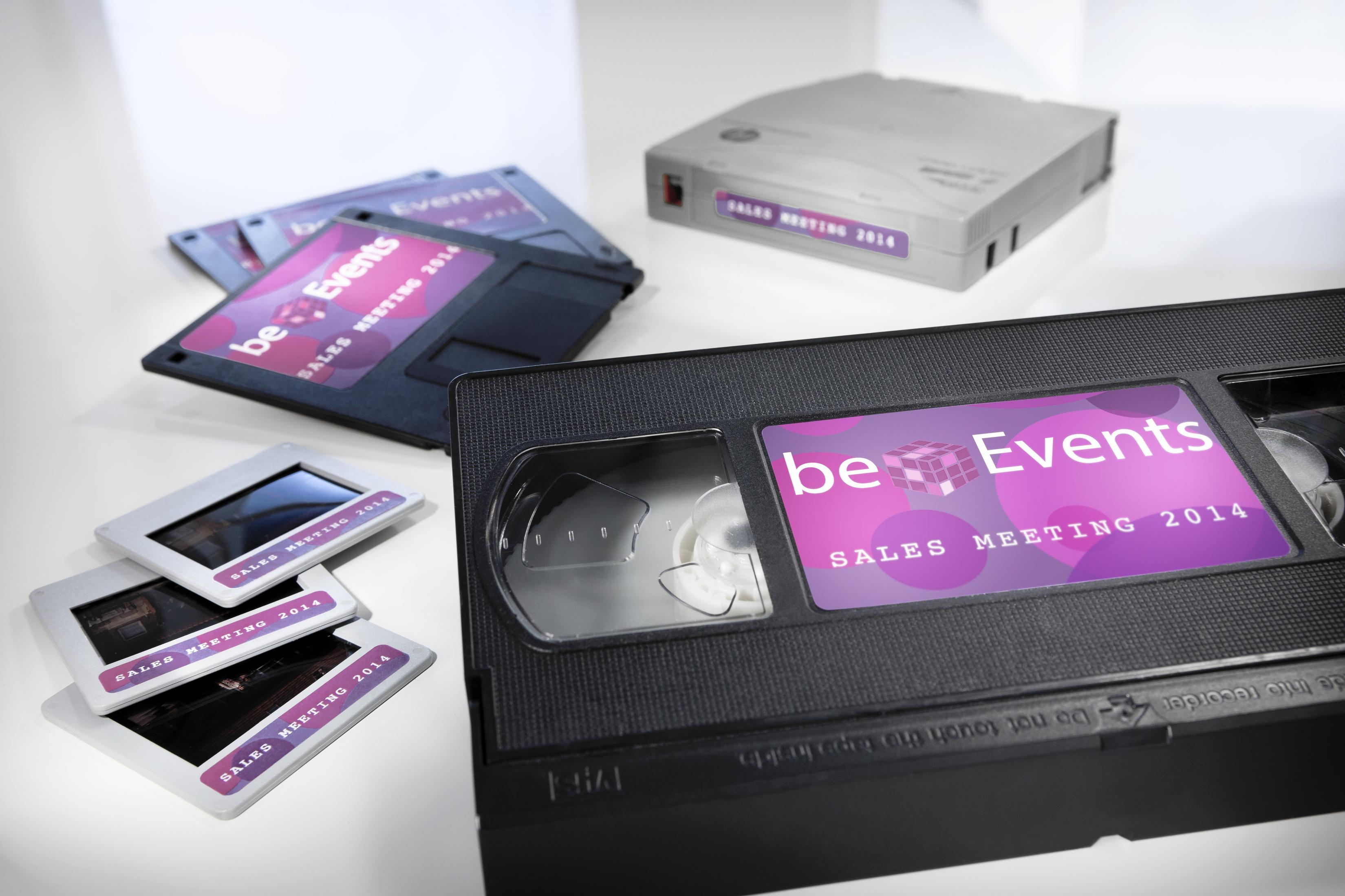 VHS-Kassette, Disketten und Dias mit der Aufschrift 'be Events Sales Meeting 2014' auf weißem Untergrund.