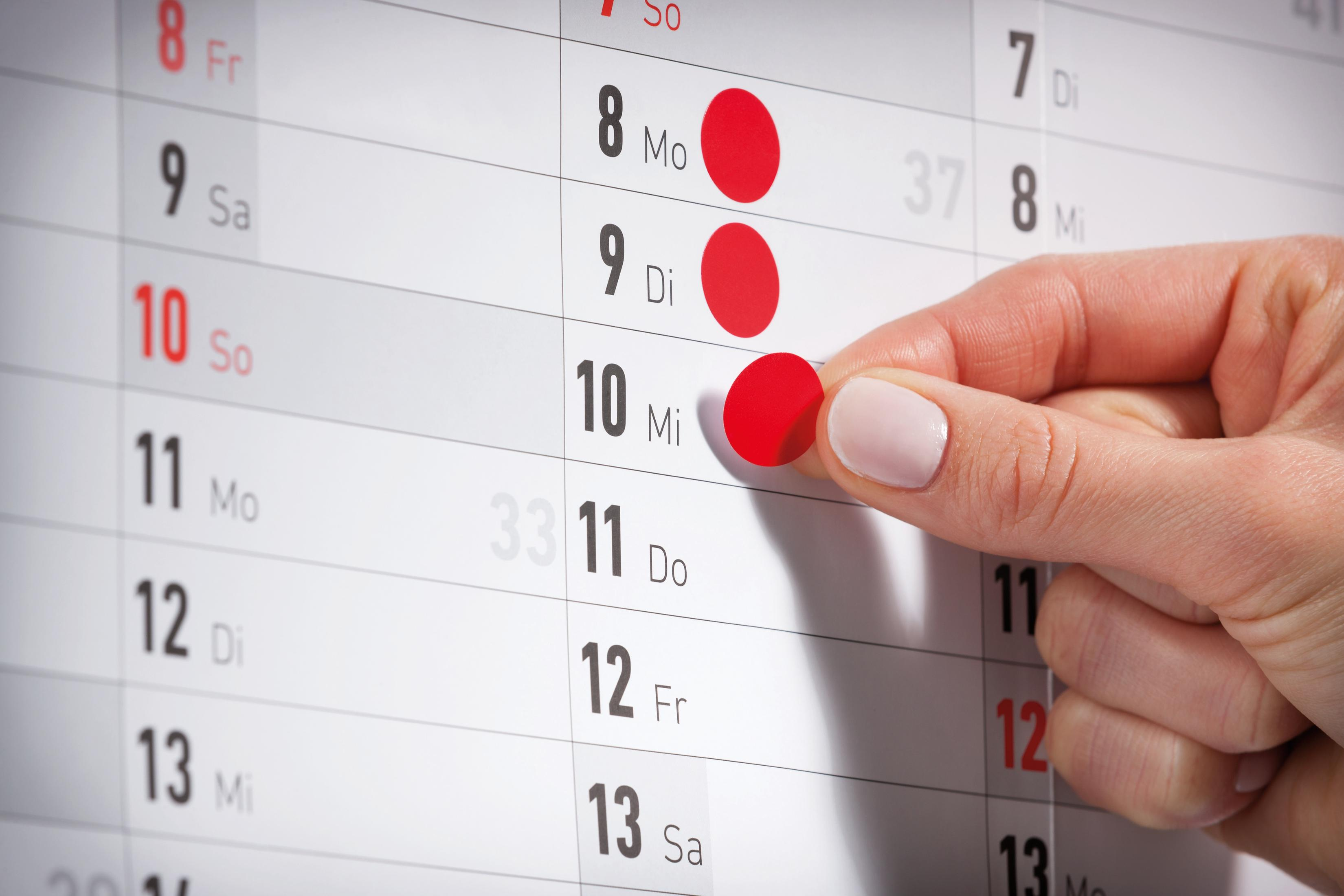 Eine Hand zeigt auf einen rot markierten Tag auf einem Kalender, der die Monatsdaten anzeigt.