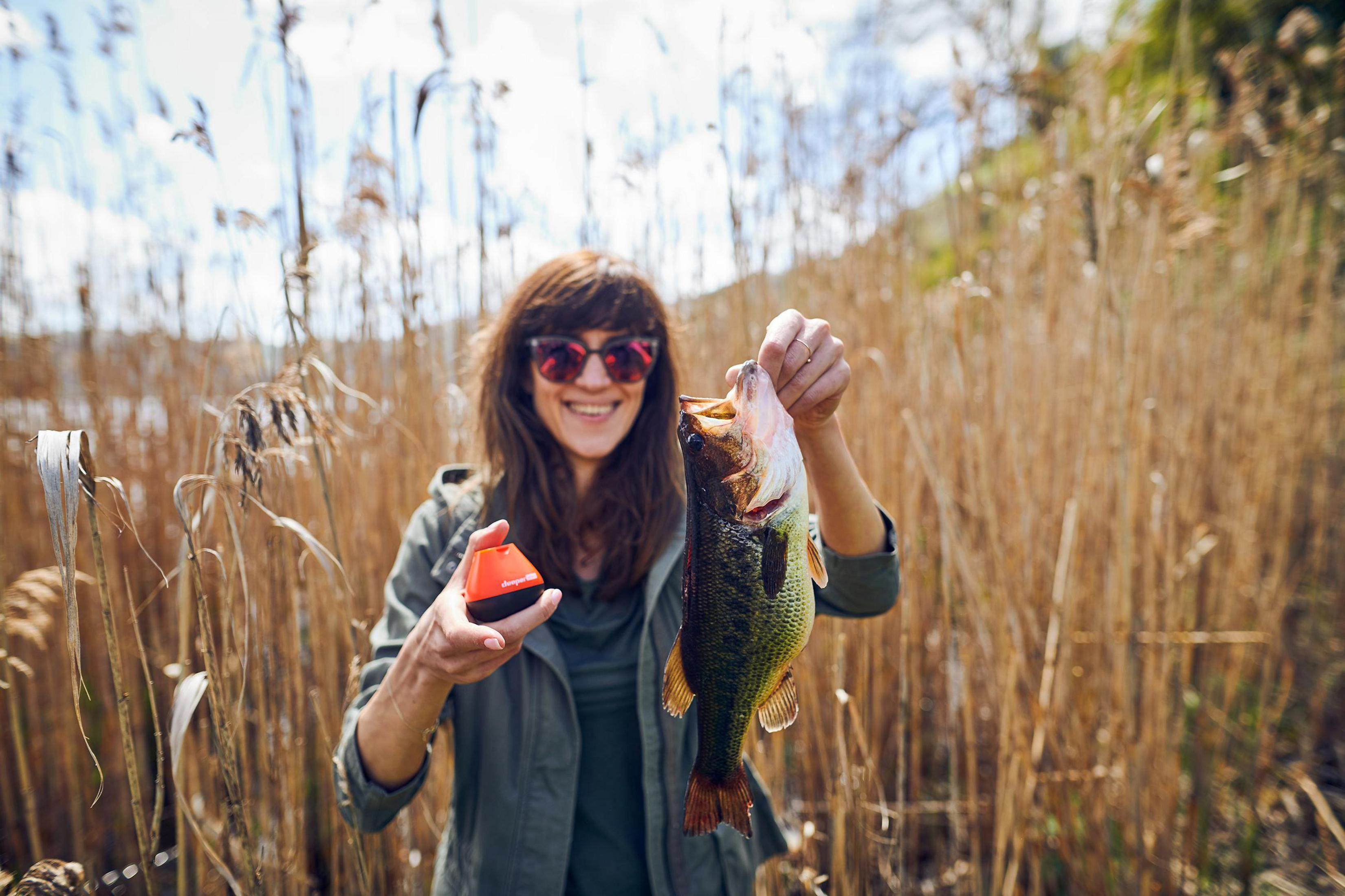 Frau hält Fisch, lächelt, trägt Sonnenbrille, steht in hohem Gras.