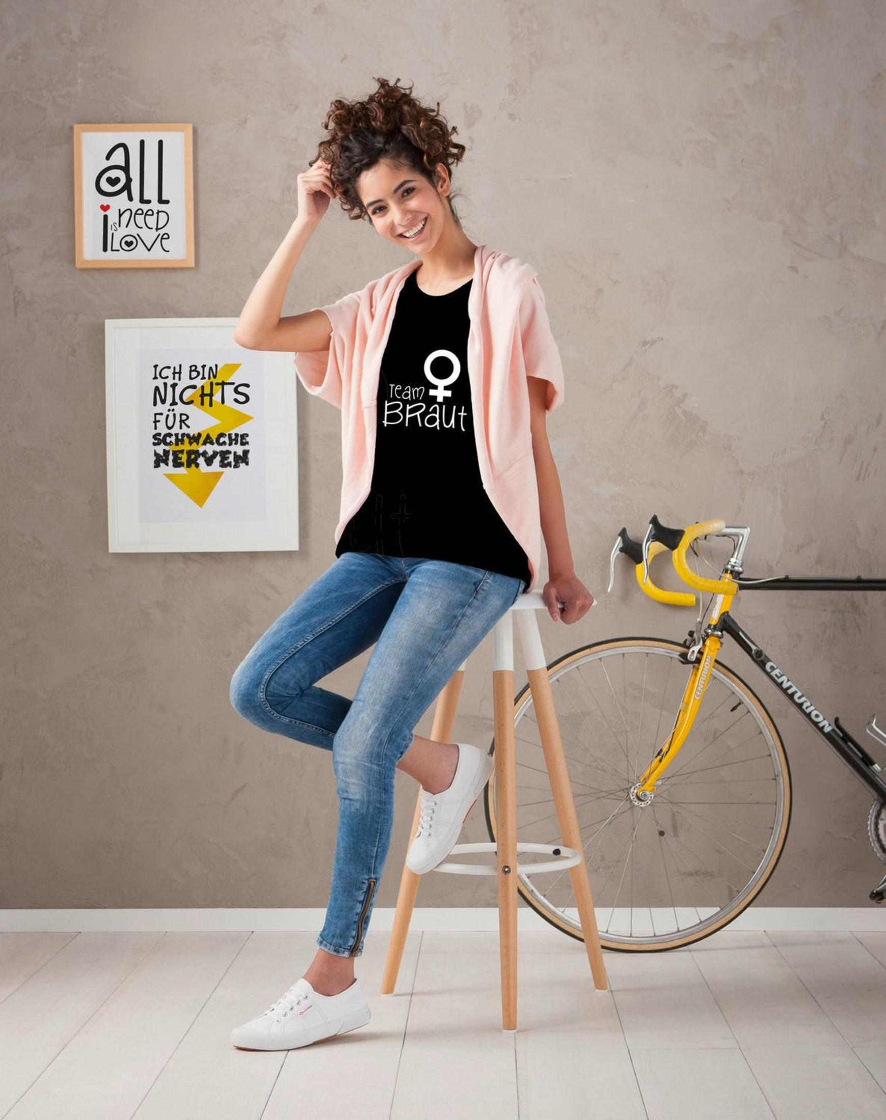 Frau in schwarzem Shirt mit 'Ich bin Braut' und Jeans, sitzt auf einem Hocker. Fahrrad im Hintergrund.
