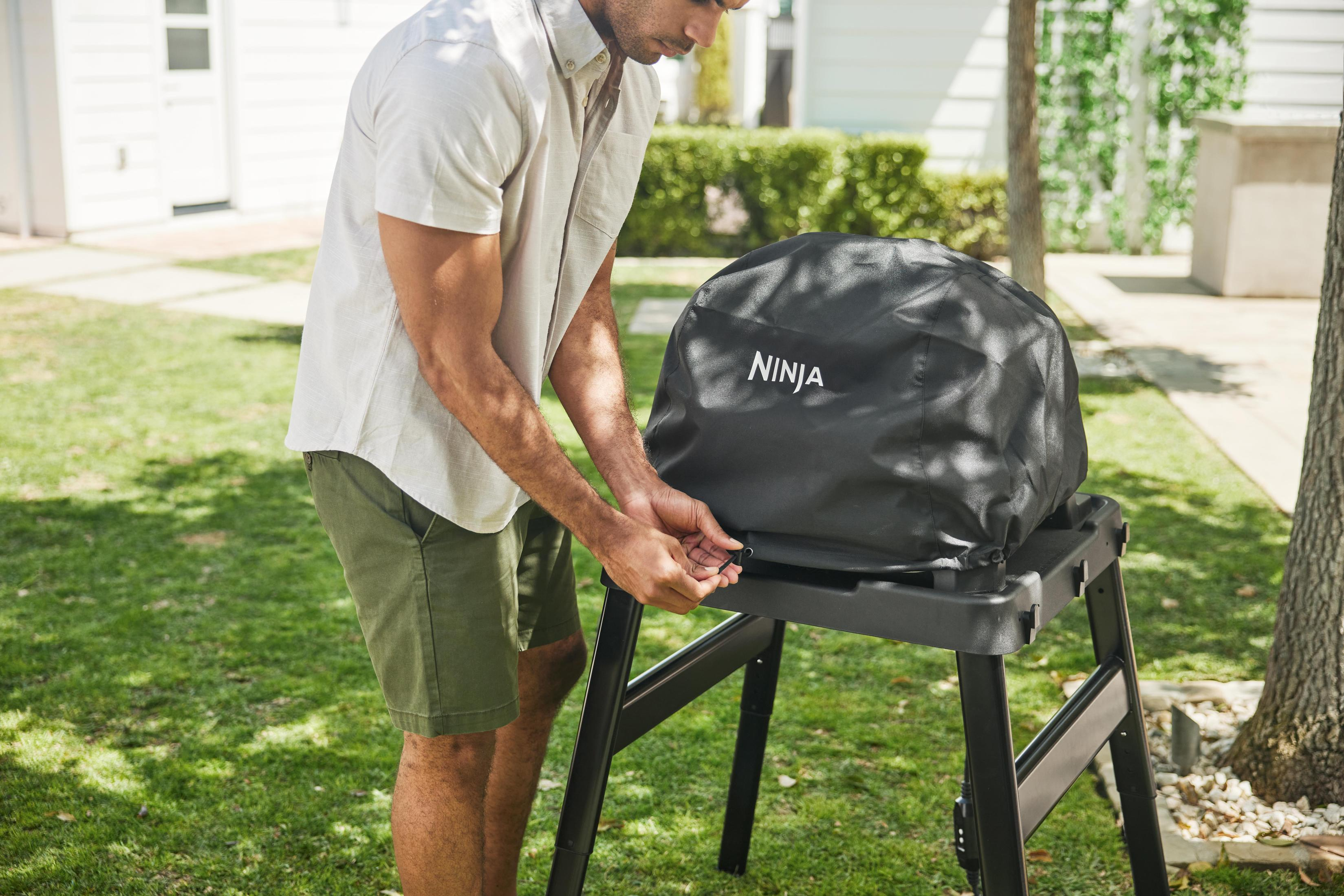 Mężczyzna w szortach przykrywa grilla czarną osłoną. Osłona ma napis NINJA.