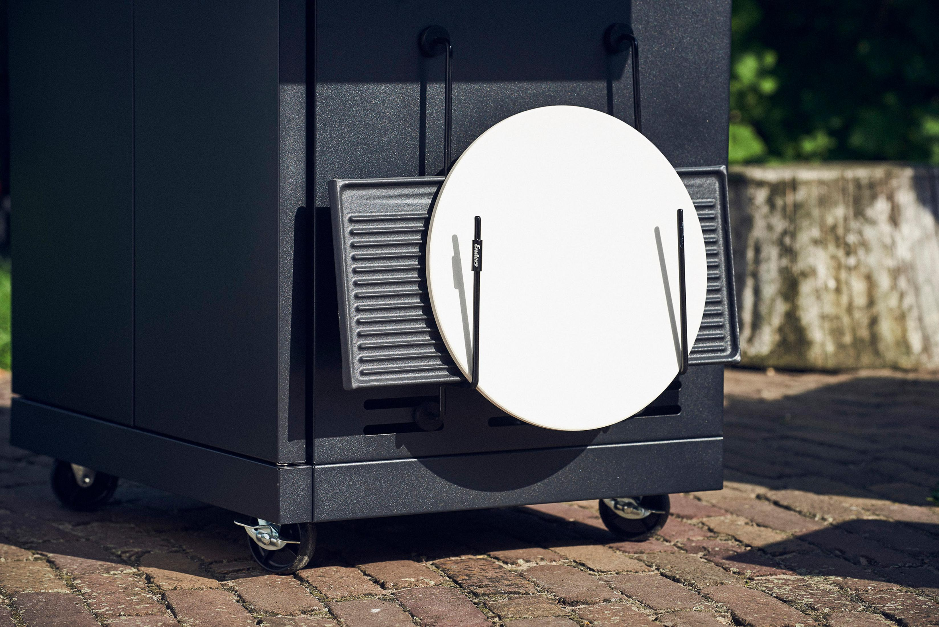 Schwarzer Outdoor-Küchenschrank mit Pizzasteinhalter und Grillrost. Ziegelboden im Hintergrund.