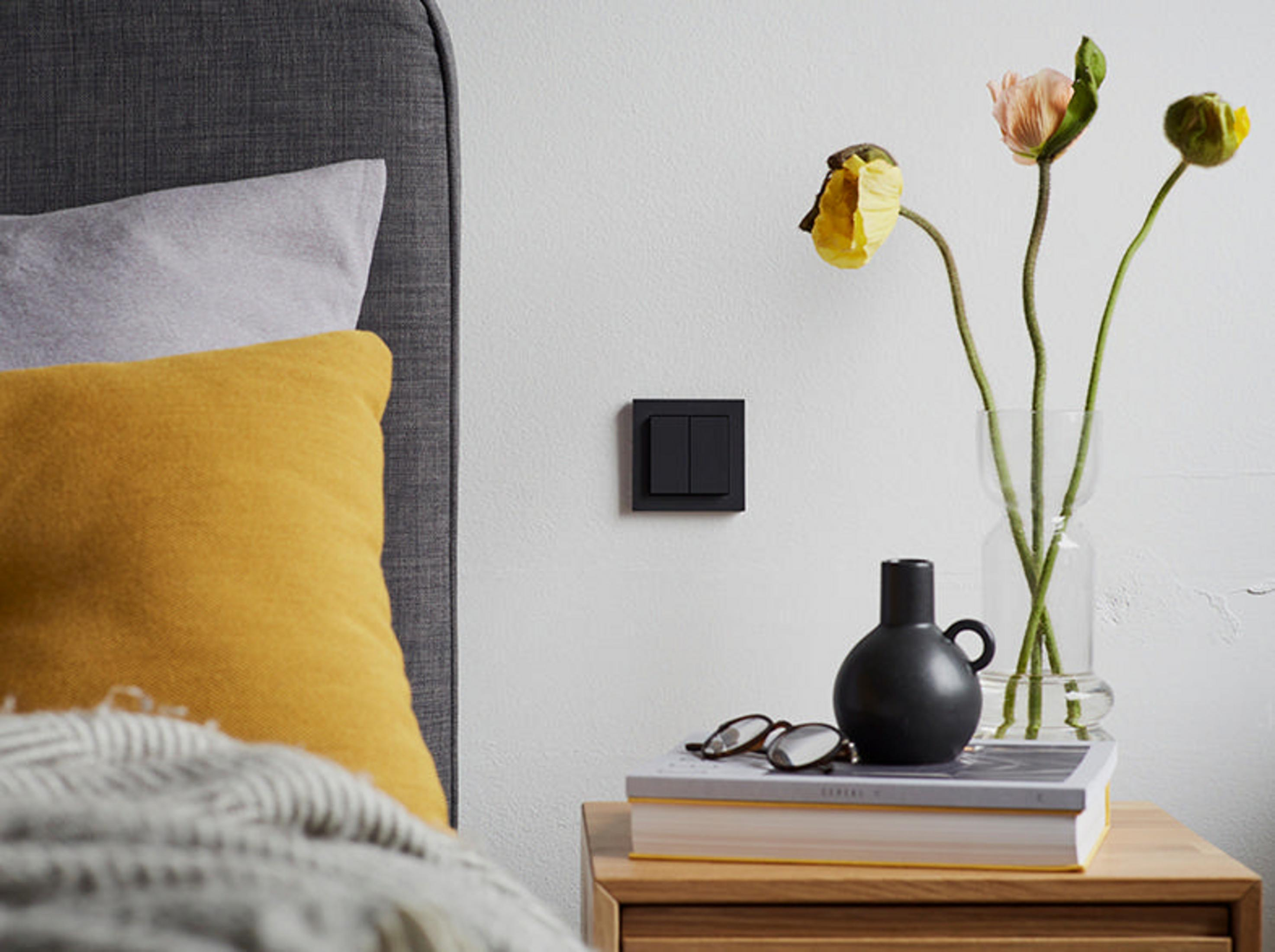 Schwarzer Lichtschalter an weißer Wand neben Nachttisch mit Blumen und schwarzer Vase.