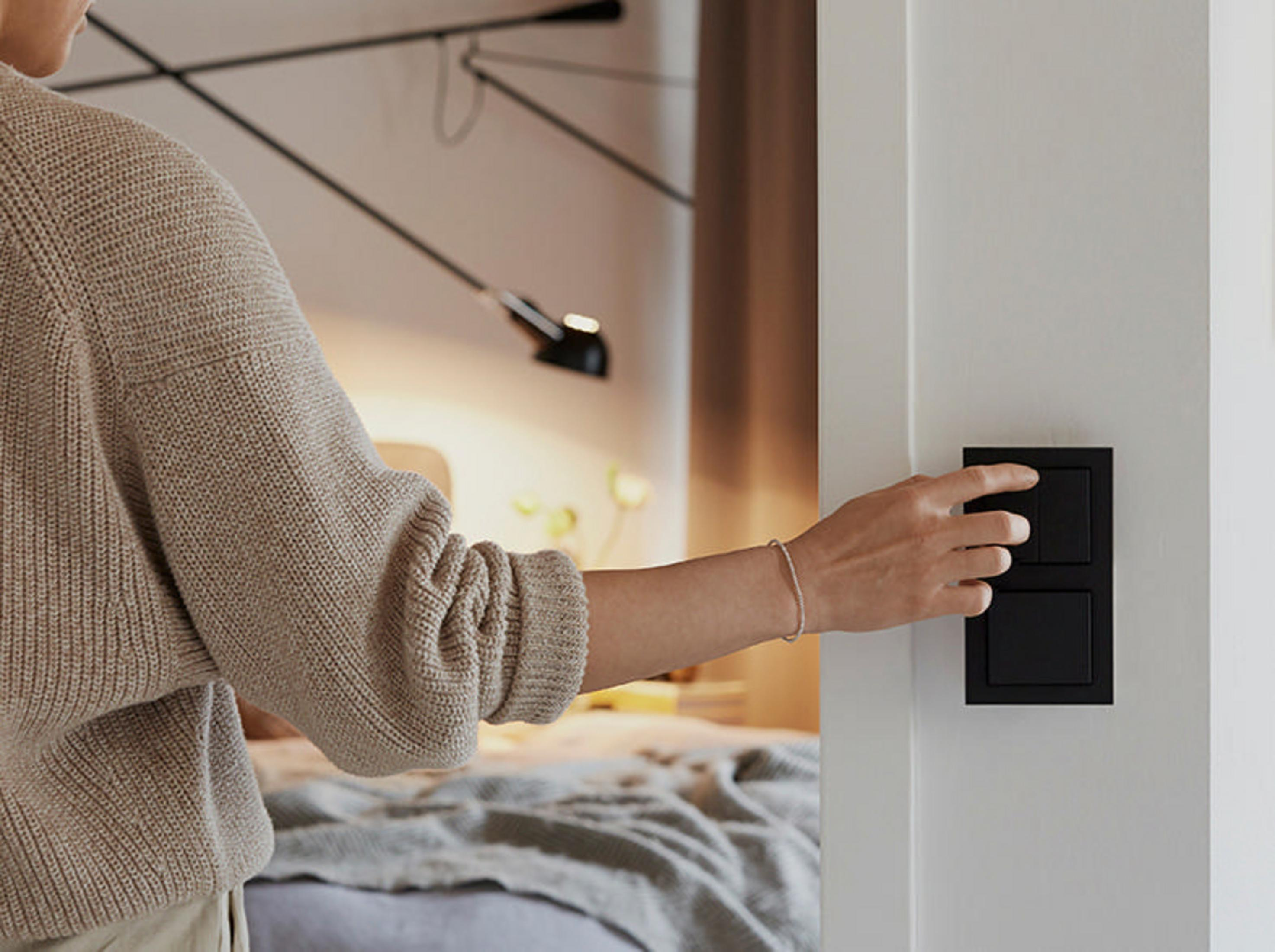 Die Hand einer Person greift nach einem schwarzen Lichtschalter an einer weißen Wand.