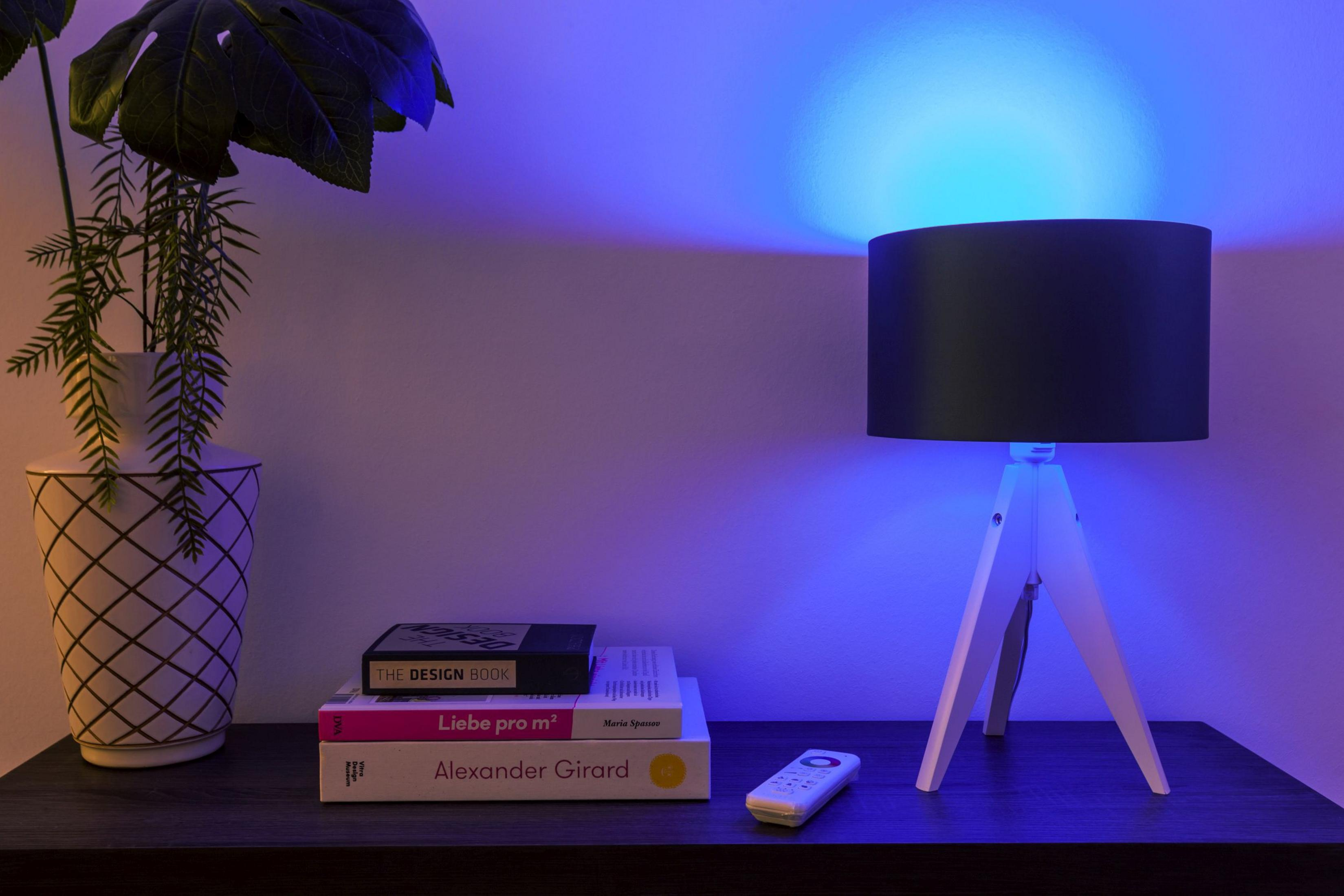 Eine Tischlampe mit schwarzem Schirm, weißem Dreibein und Büchern auf einem Tisch, blau beleuchtet.