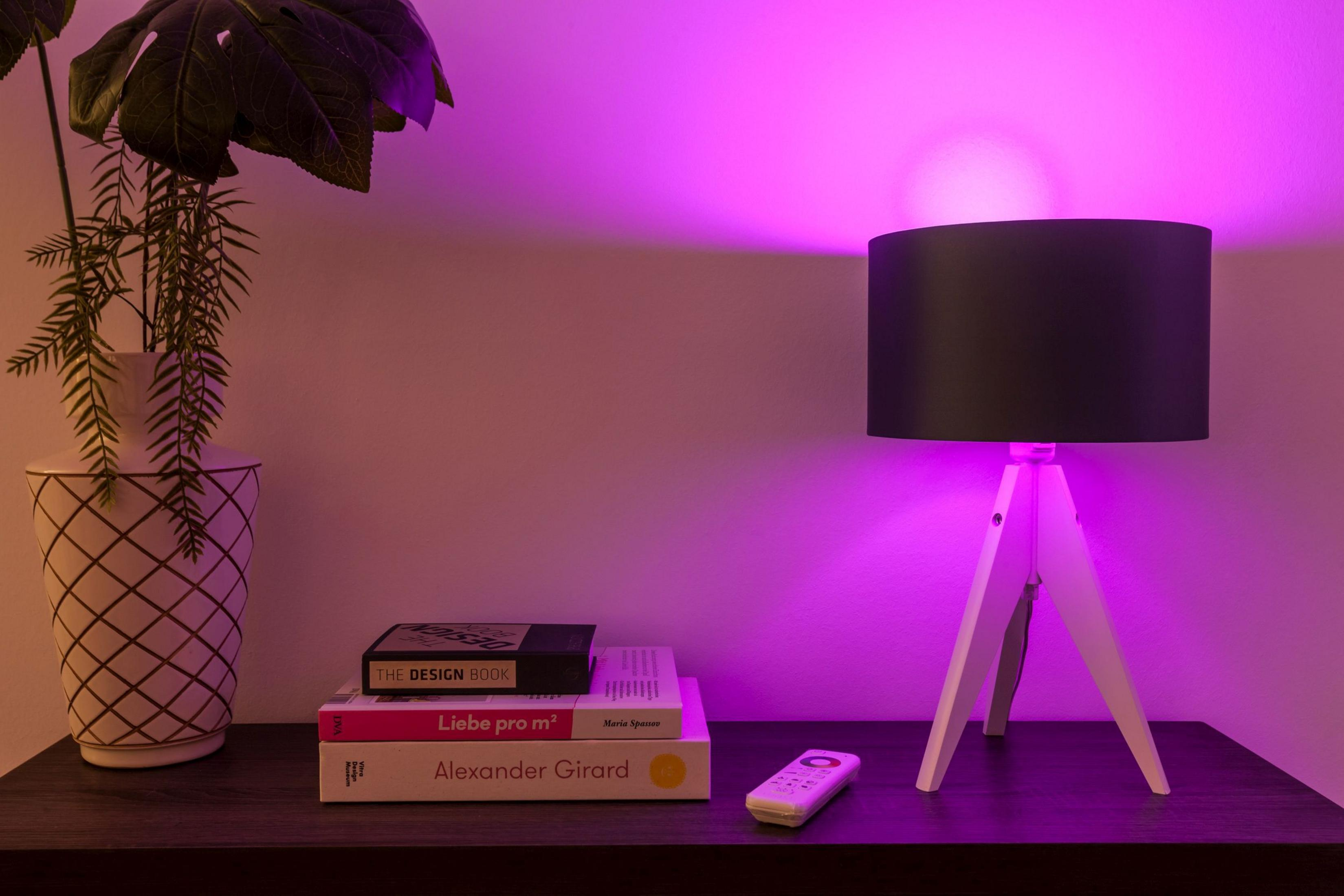 Eine Lampe mit schwarzem Schirm, weißem Dreibein, Büchern auf einem Tisch, pink beleuchtet.