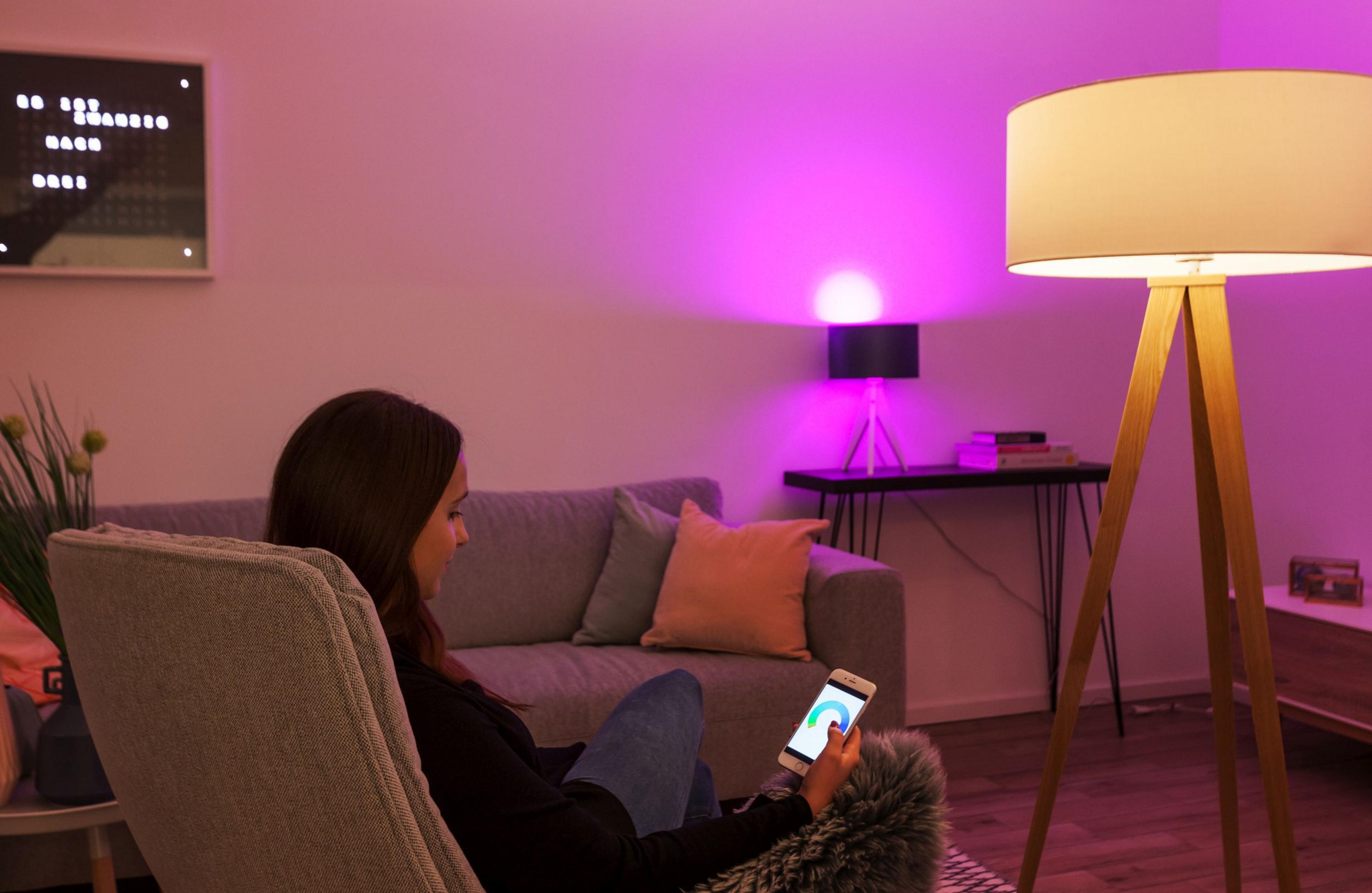 Eine Frau benutzt ein Smartphone, in der Nähe eines Sofas und einer Stehlampe, alles rosa beleuchtet.