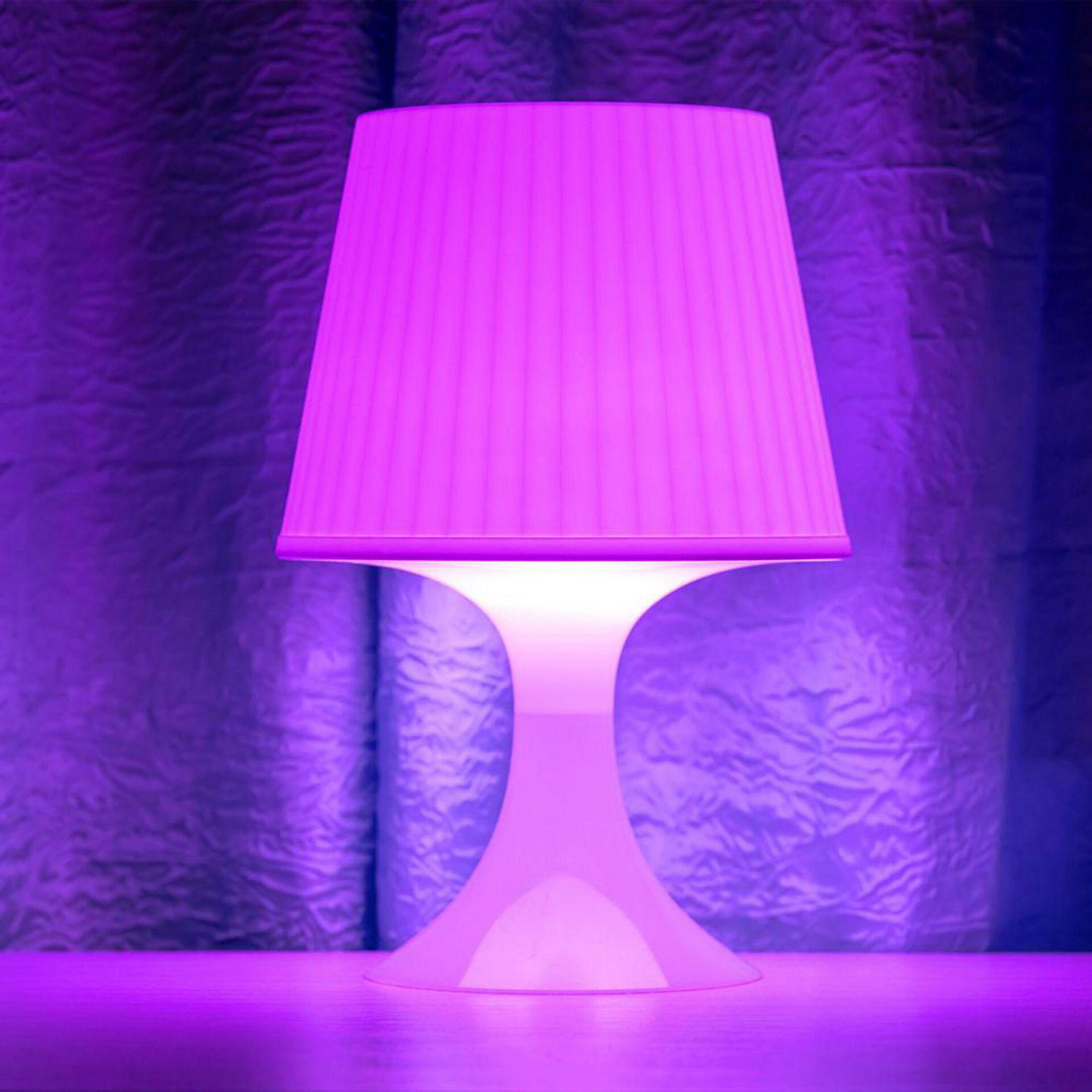 Eine weiße Lampe mit rosa Lampenschirm, die Lampe steht auf einer Oberfläche, der Hintergrund ist dunkelblau.