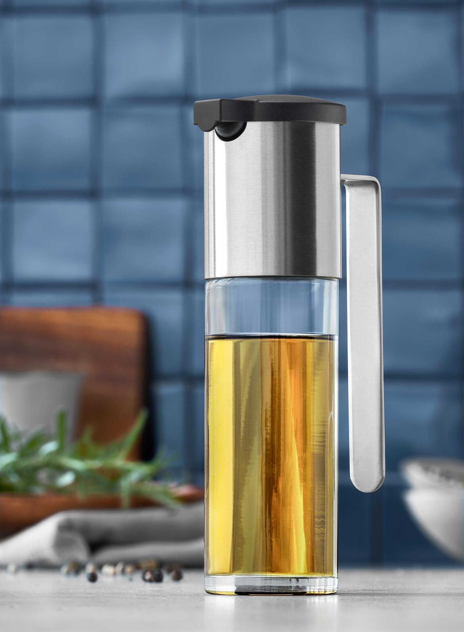 Ein Glas-Ölspender mit Olivenöl, vor blauem Fliesen-Hintergrund.