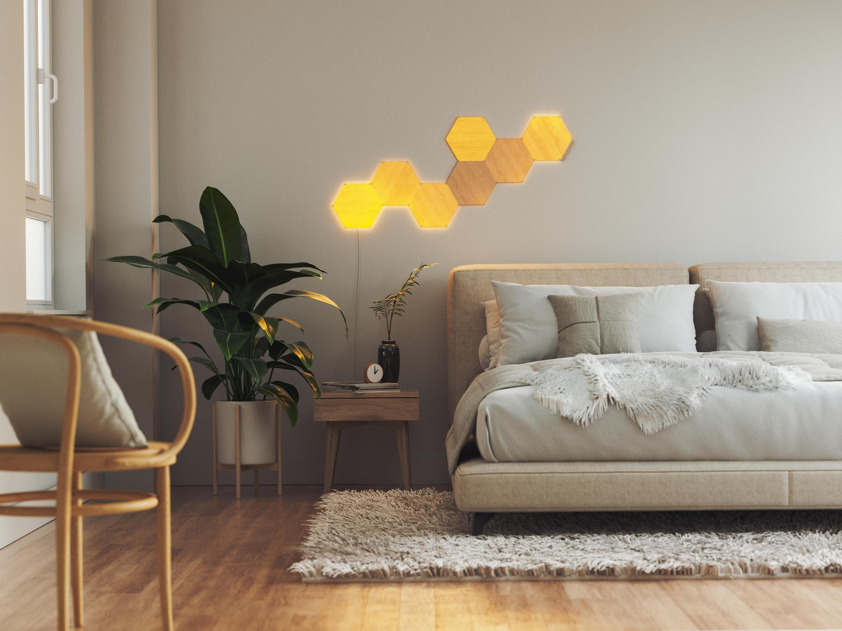 Scena di camera da letto con luci a parete esagonali in legno, letto, pianta e sedia. Luce calda.
