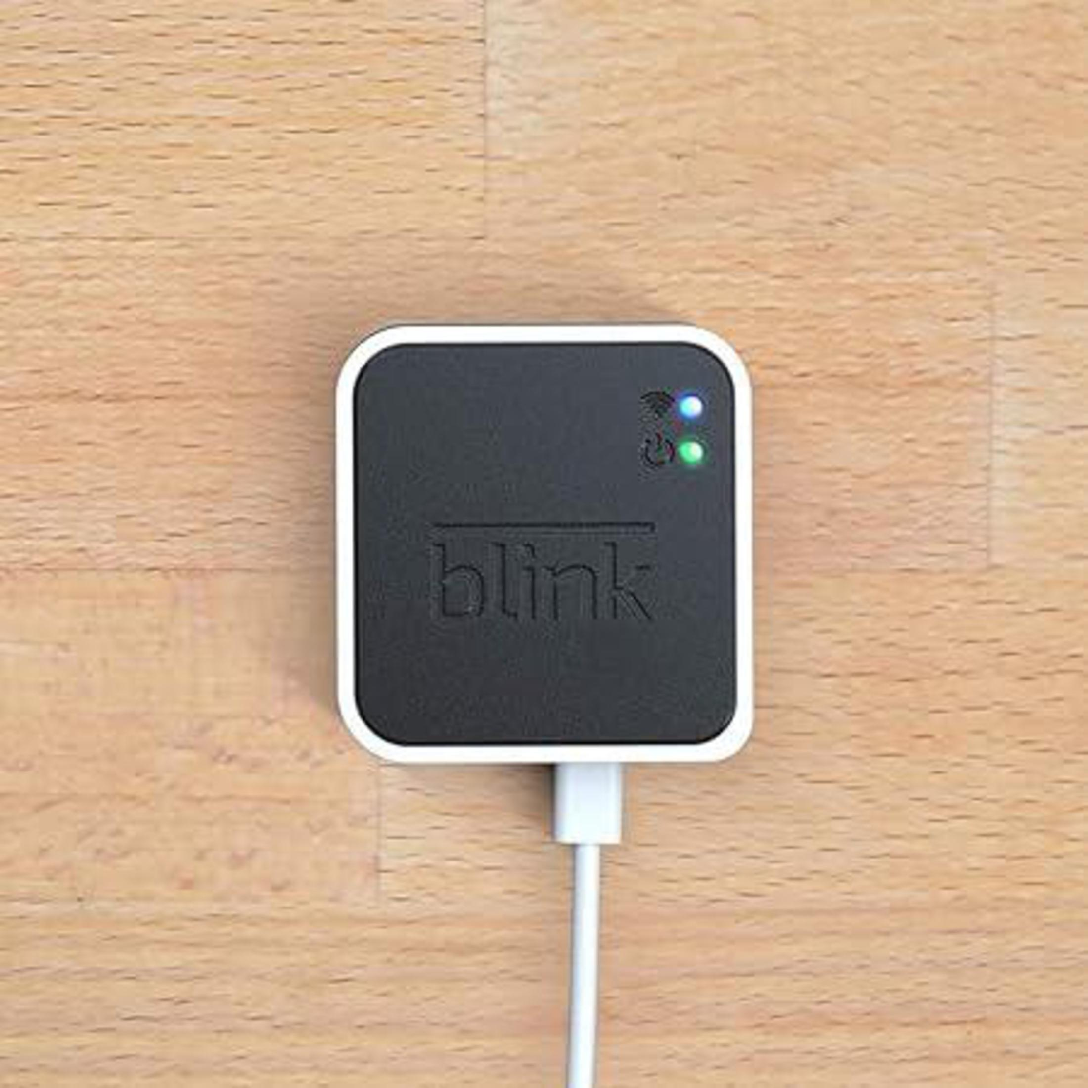 Schwarzes quadratisches Gerät mit weißem Rand, weißes Kabel auf Holzoberfläche. Hat das Wort 'blink' und Lichter.