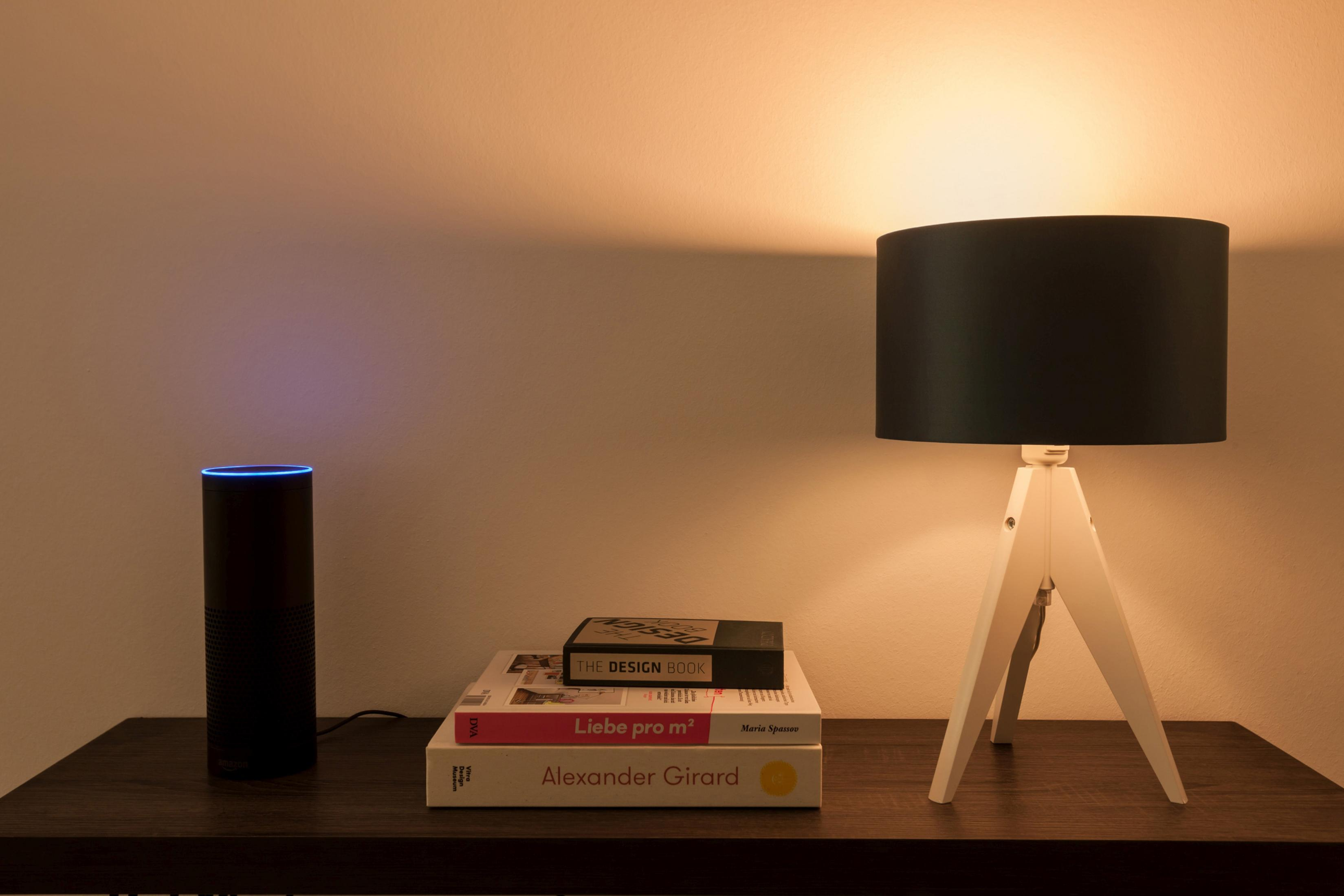 Eine Raumszene mit einer Lampe, Büchern und einem Smart Speaker auf einem Tisch. Die Lampe wirft warmes Licht.