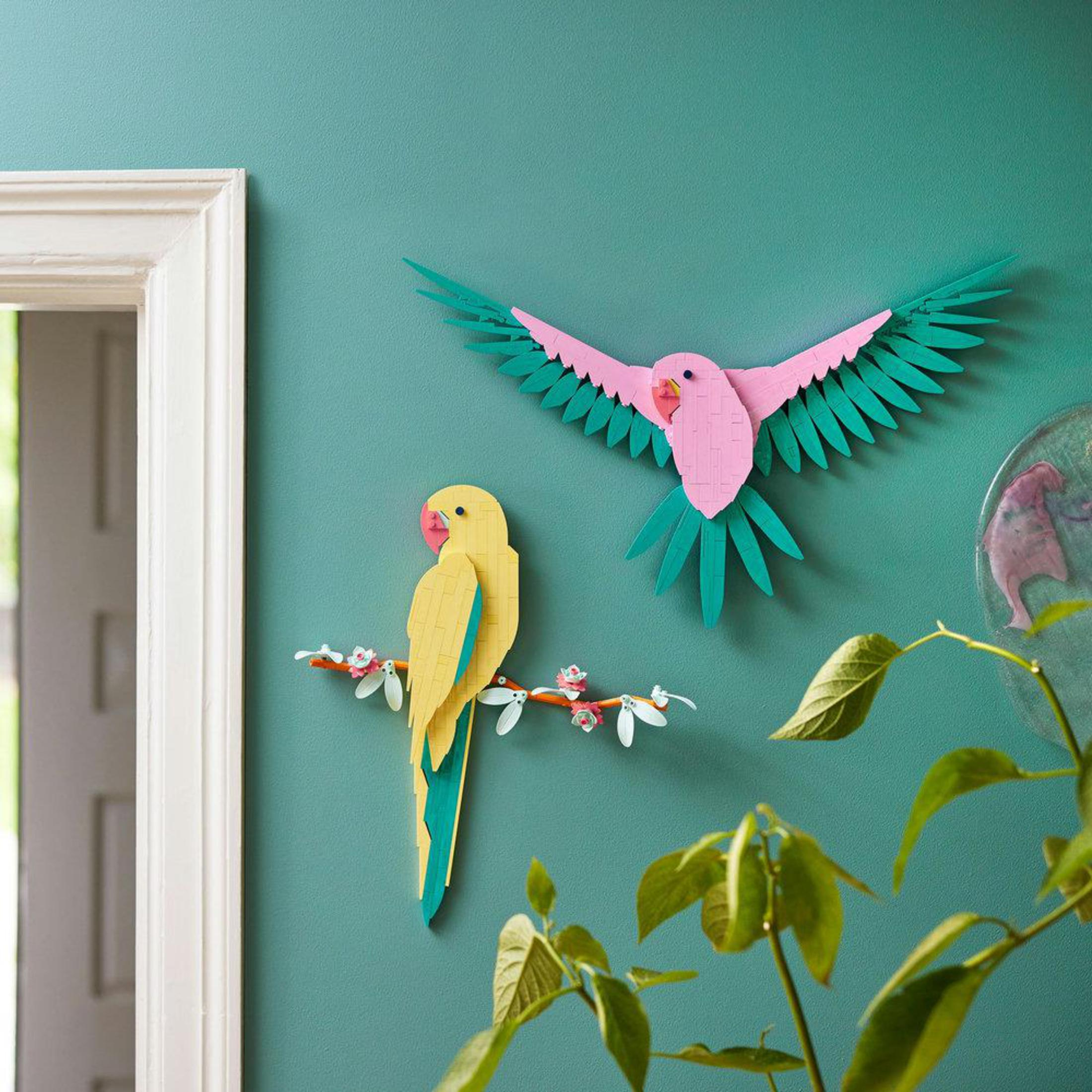 Twee papieren papegaaien op een turquoise muur. Eén vliegt, de ander zit op een tak.