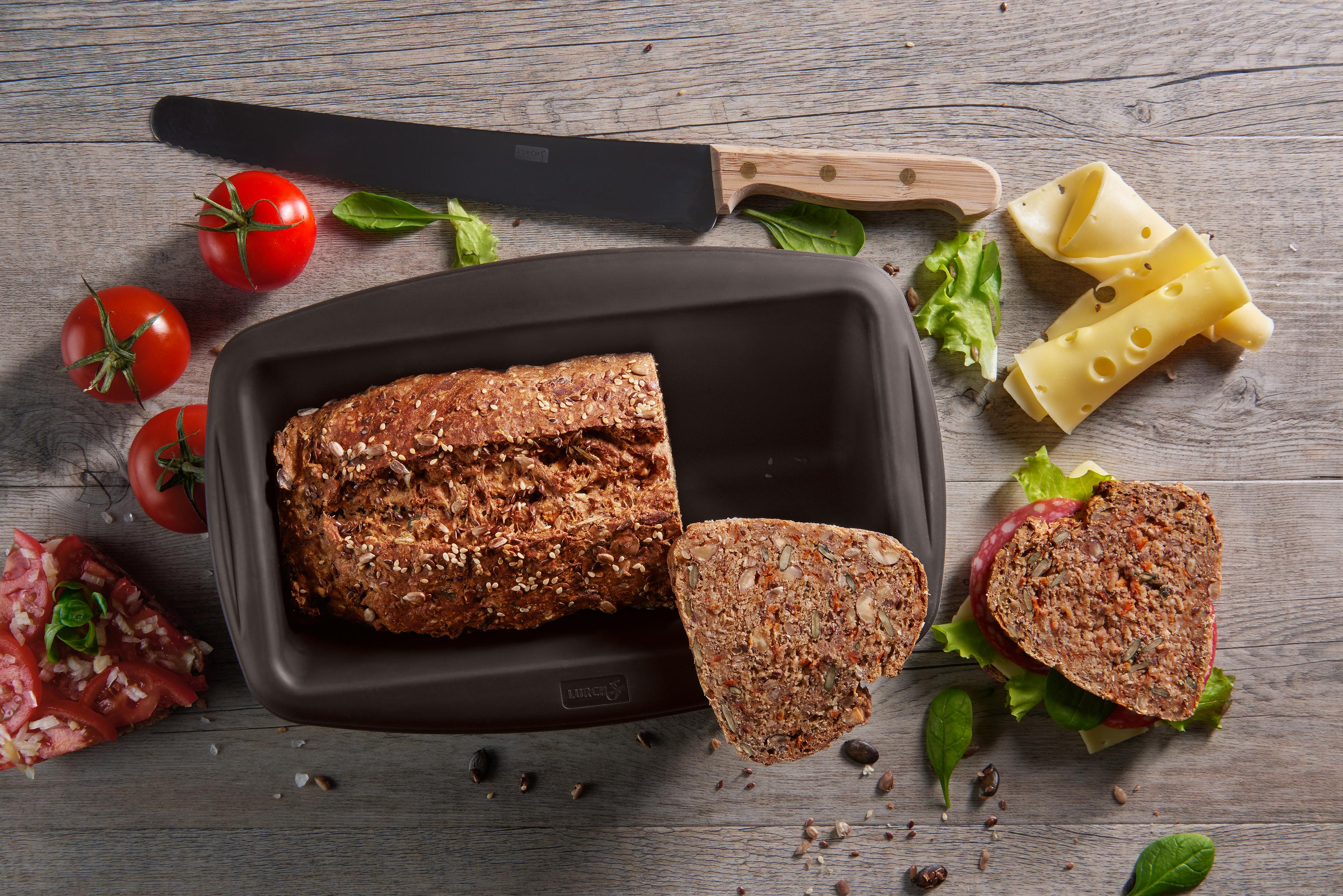 Ein Brotlaib in einer braunen Form, geschnittenes Brot, Tomaten, Käse und Messer auf Holz.