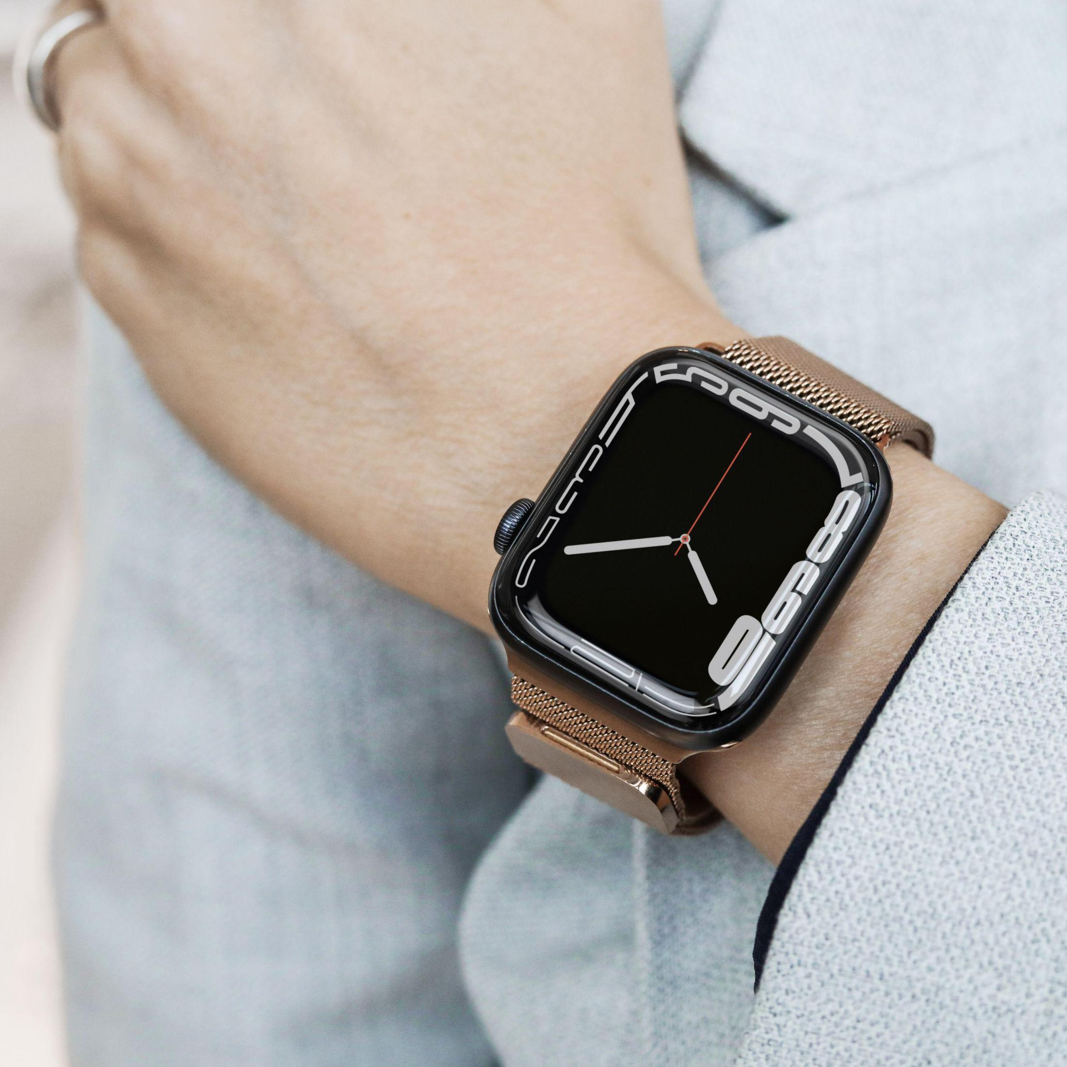Ein Handgelenk trägt eine silberne Apple Watch mit einem goldenen Mailänder Armband vor einer grauen Jacke.