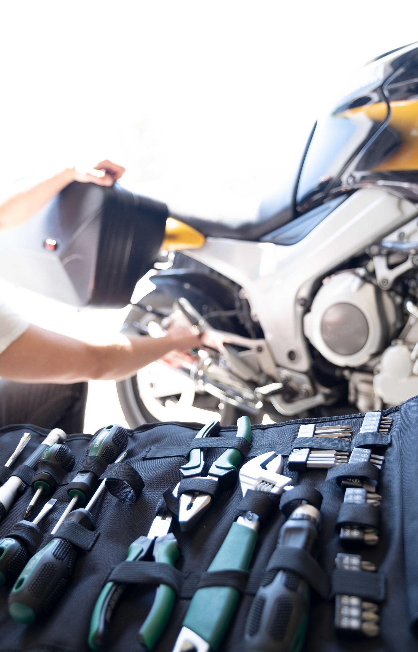 Werkzeuge in einer schwarzen Tasche ausgebreitet, mit einem Motorrad und einer Person im Hintergrund.
