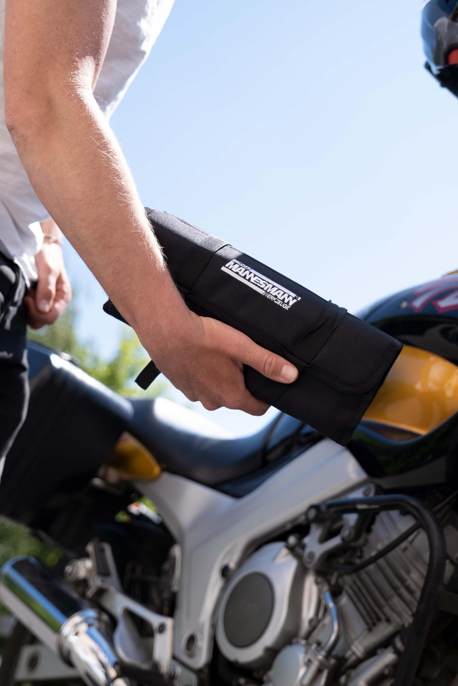 Eine Person hält eine schwarze Werkzeugrolle vor einem Motorrad. Die Rolle hat ein weißes Logo.