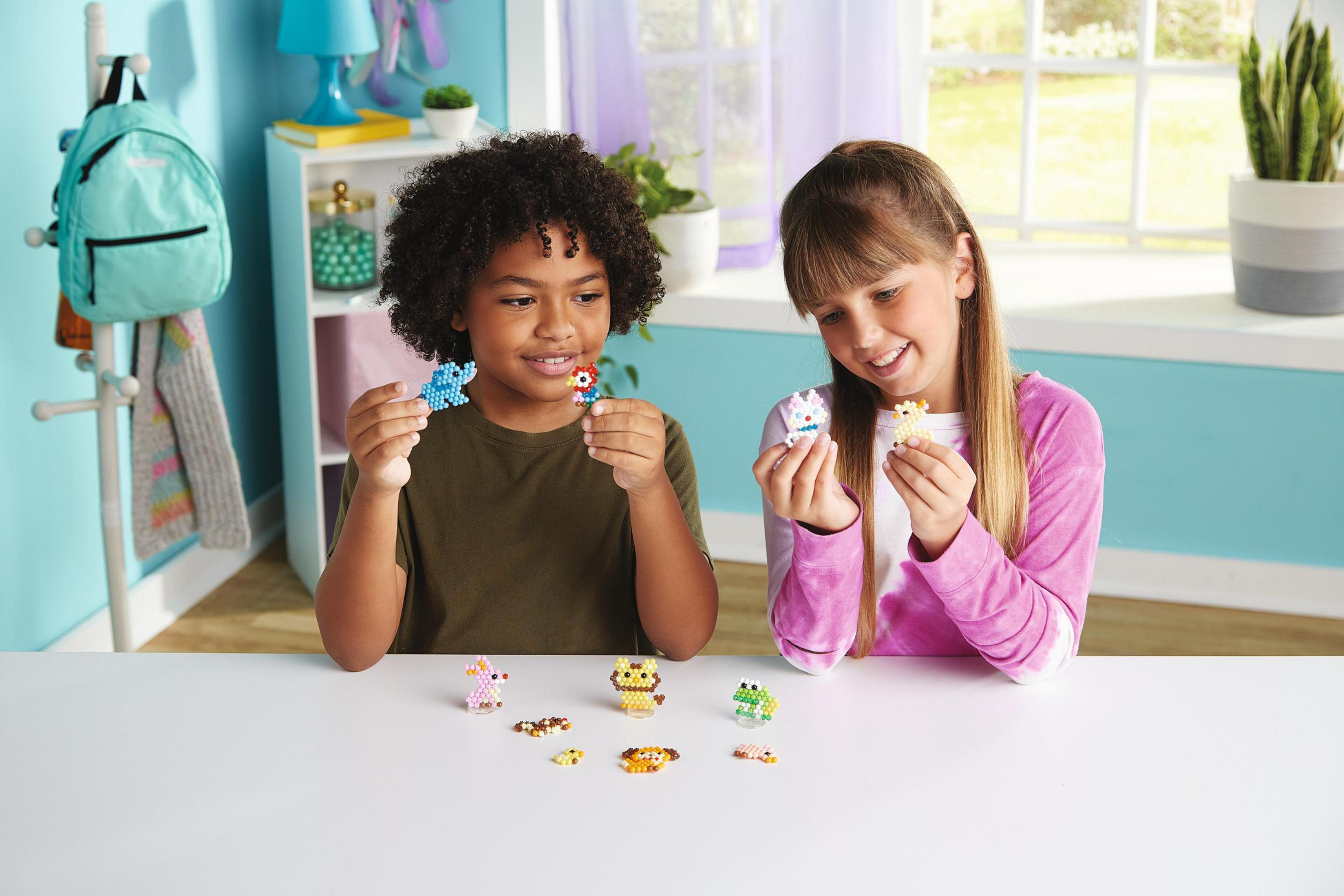 Zwei Kinder sitzen an einem Tisch. Sie halten kleine, aufgefädelte Tierfiguren. Andere Figuren liegen auf dem Tisch.