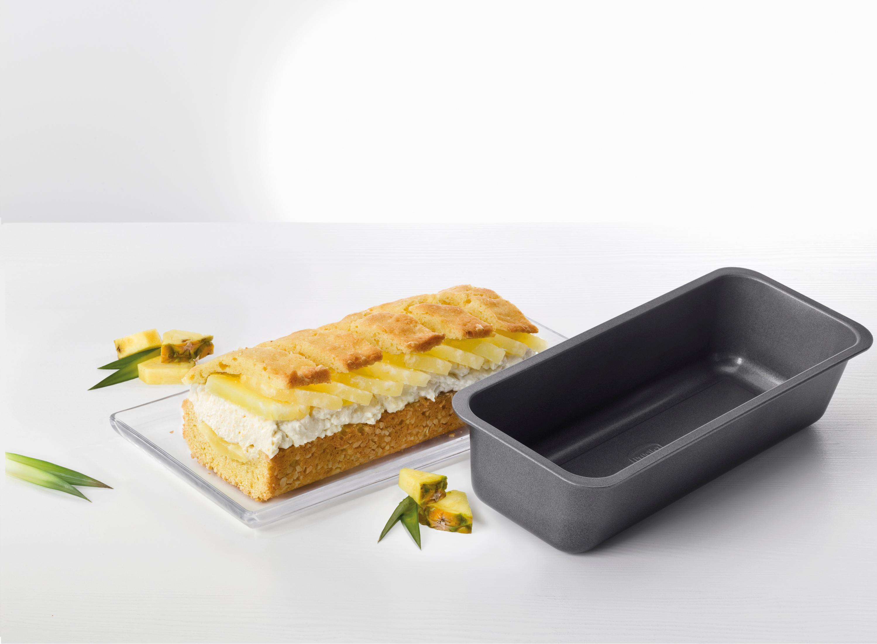 Ein Kuchen auf einem Glasteller, Ananasscheiben und eine Backform auf einer weißen Oberfläche. Der Kuchen ist mit Sahne gefüllt.