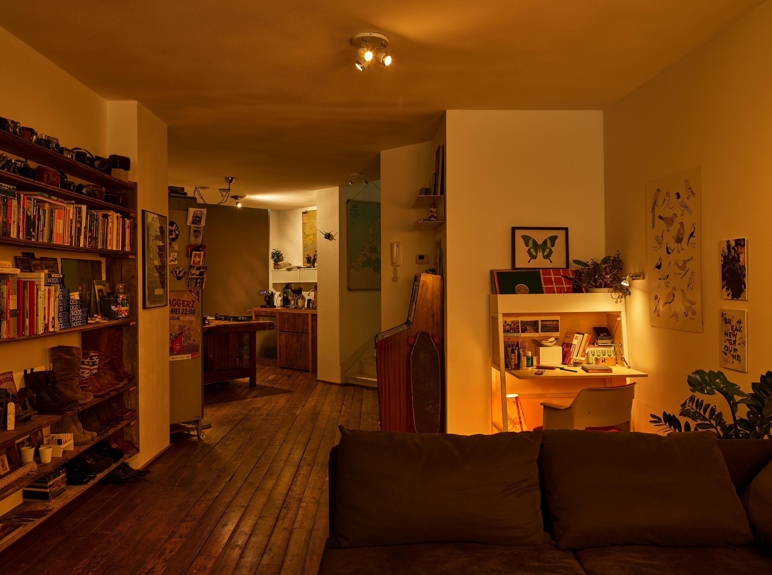 Ein warm beleuchtetes Wohnzimmer mit Büchern, Kunst und einem Schreibtisch. Eine Tür führt in die Küche.