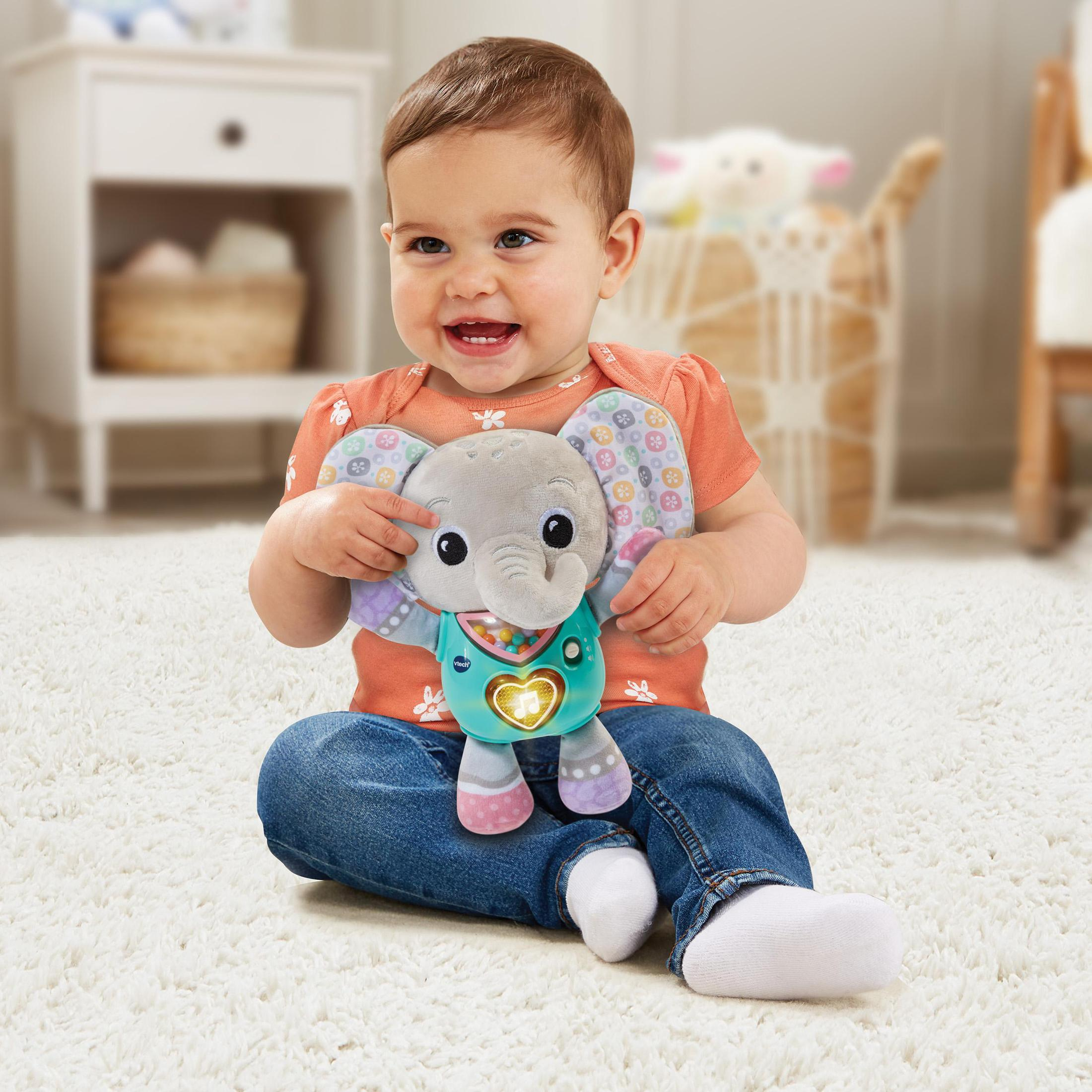 Baby lächelt, hält graues Elefantenspielzeug. Trägt orangefarbenes Hemd und Jeans, sitzt auf einem Teppich.