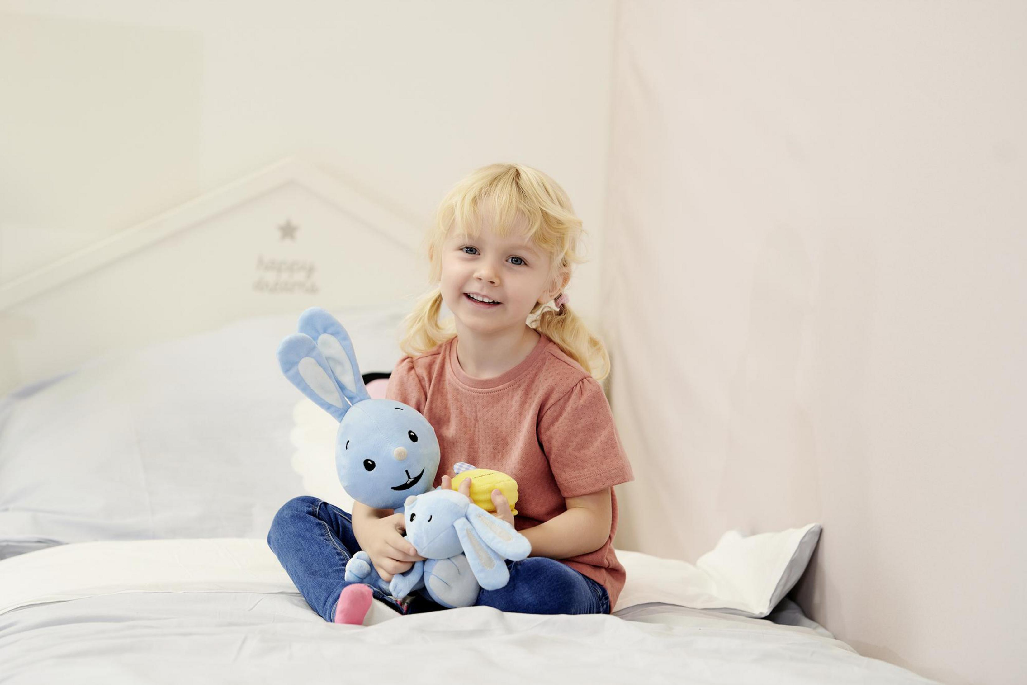 Ein lächelndes Mädchen, das zwei blaue Plüschhasen hält und auf einem Bett sitzt.