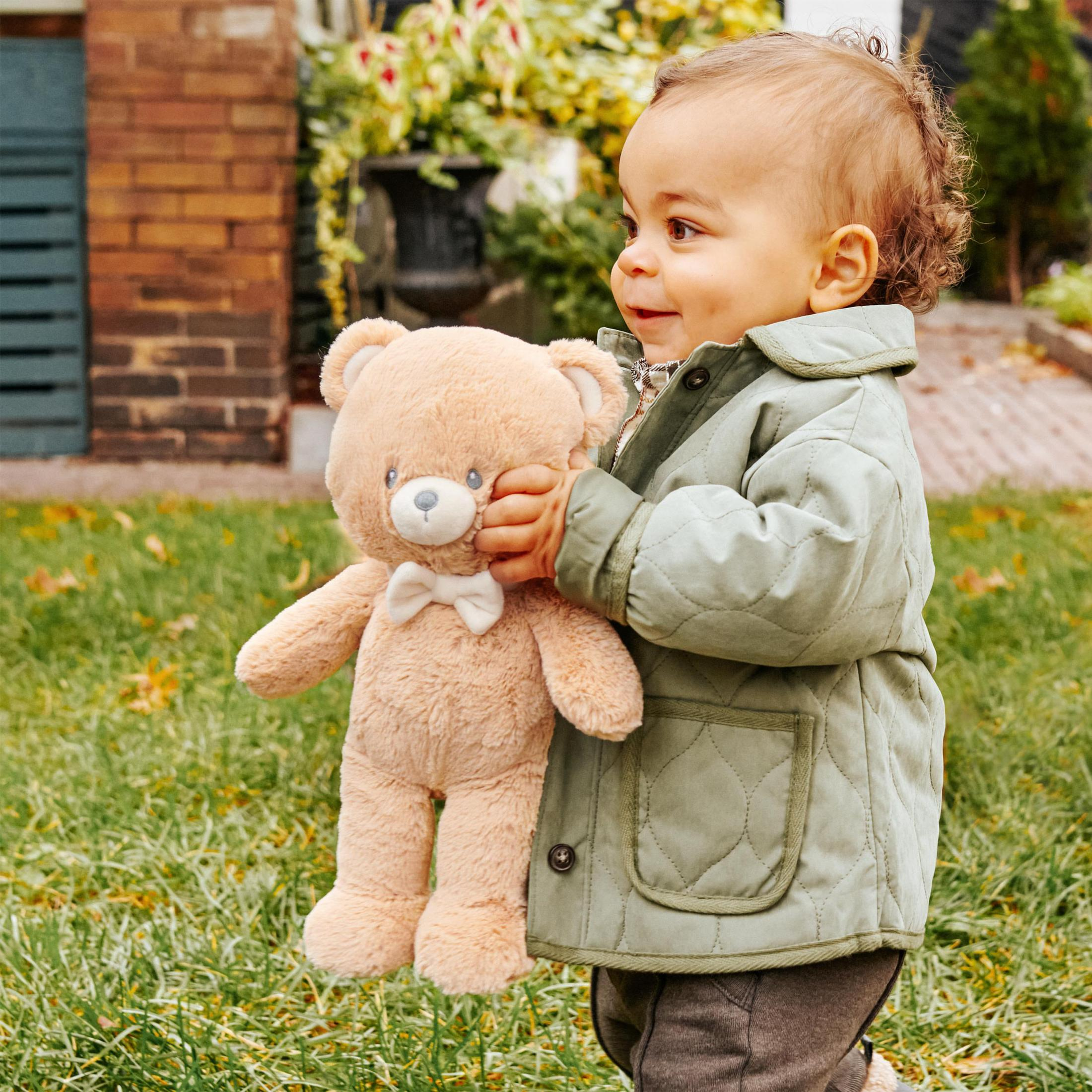 Kleinkind hält einen Teddybären, trägt eine grüne Jacke, steht draußen.